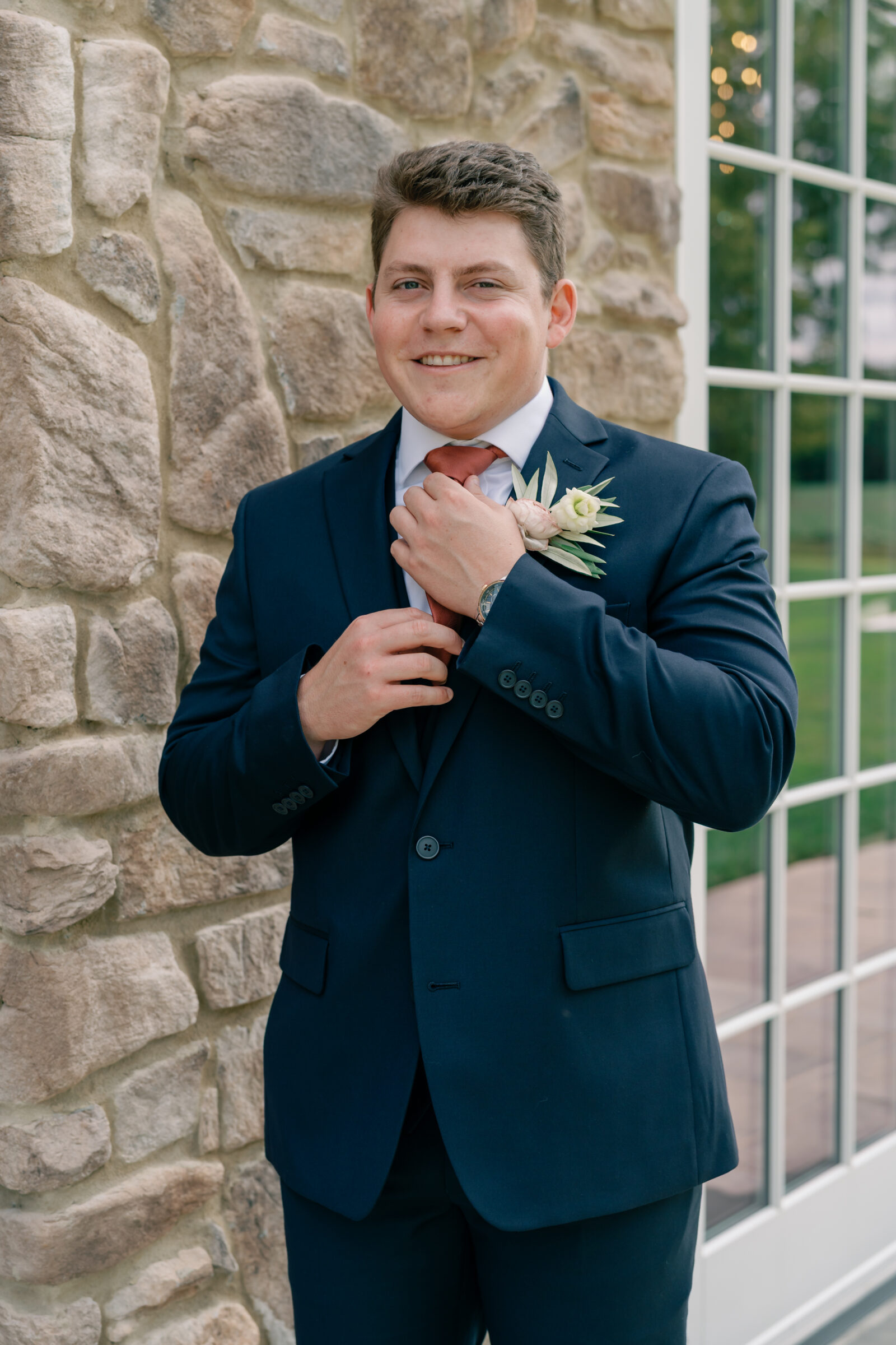 Groom in a navy suit adjusting his tie with a boutonniere, smiling outside the stone manor at Burlington Weddings & Events in Charles City VA.