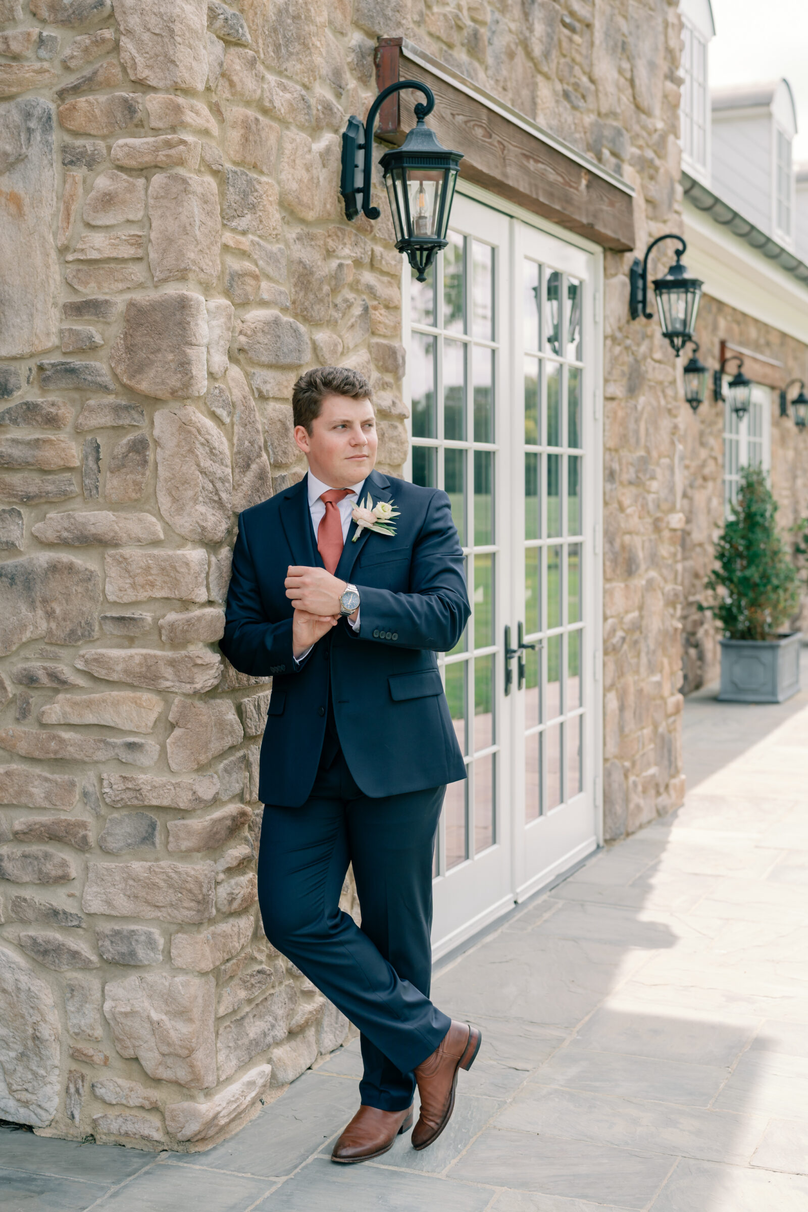 Groom in a navy suit with a blush boutonniere leaning against the stone exterior of Burlington Weddings & Events in Charles City VA.
