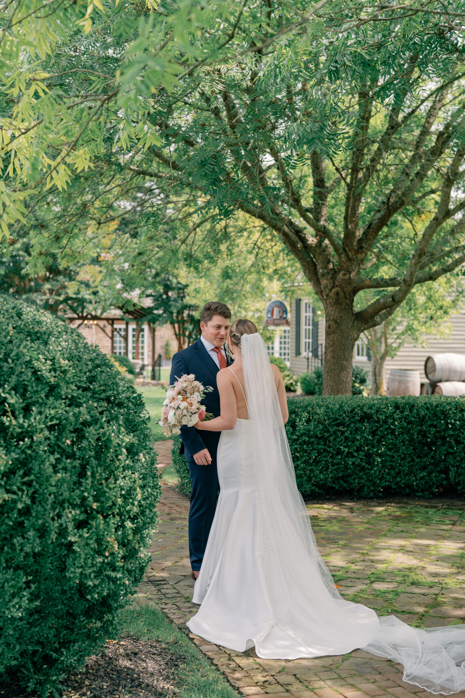 Bride in a fitted satin gown with a long veil holding a pastel bouquet during a first look with the groom in the garden at Burlington Weddings & Events in Charles City VA.