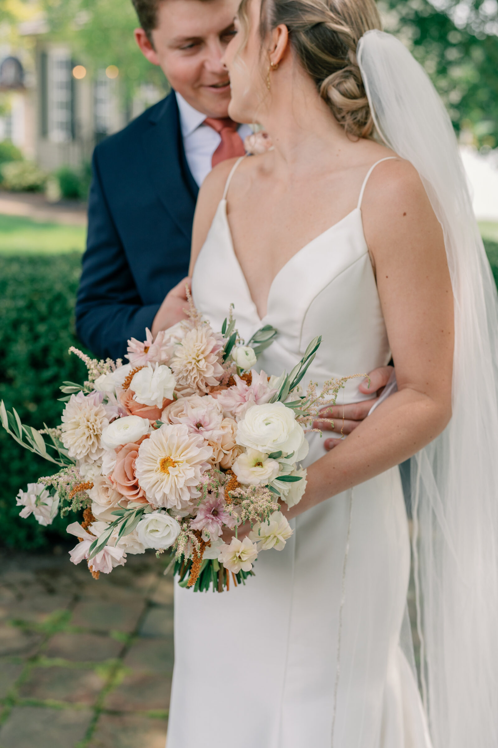 Romantic close-up of bride holding a pastel bouquet of dahlias, roses, and ranunculus while sharing a tender moment with the groom at Burlington Weddings & Events in Charles City VA.