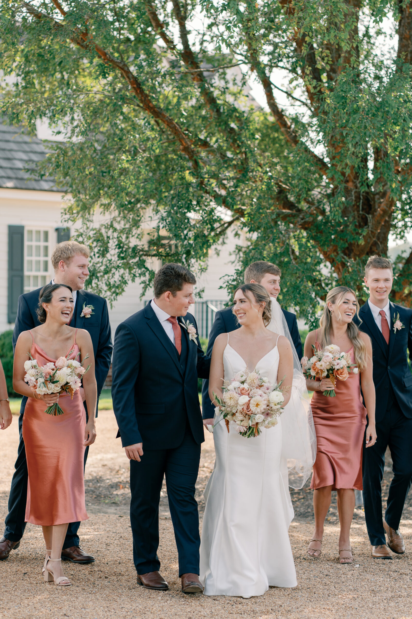 Bride and groom walking with bridesmaids in satin rust dresses and groomsmen in navy suits, laughing together outside Burlington Weddings & Events in Charles City VA.
