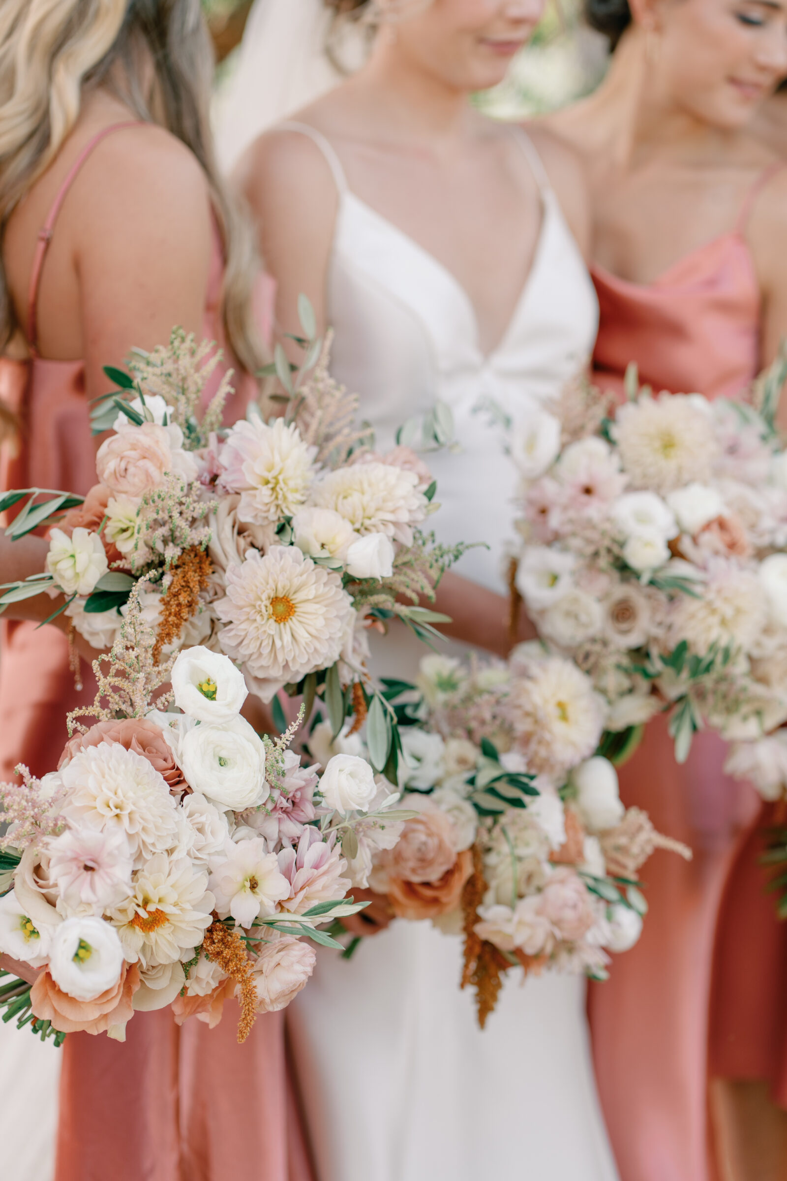 Close-up of bride and bridesmaids holding pastel bouquets of dahlias, roses, and ranunculus with satin rust dresses.