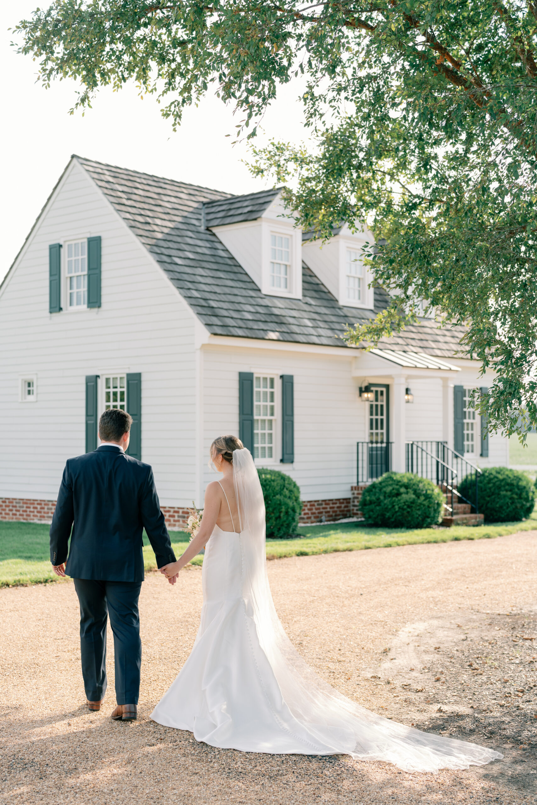 Bride in a satin gown with a long veil holding hands with the groom as they walk together outside the white manor house at Burlington Weddings & Events in Charles City VA.