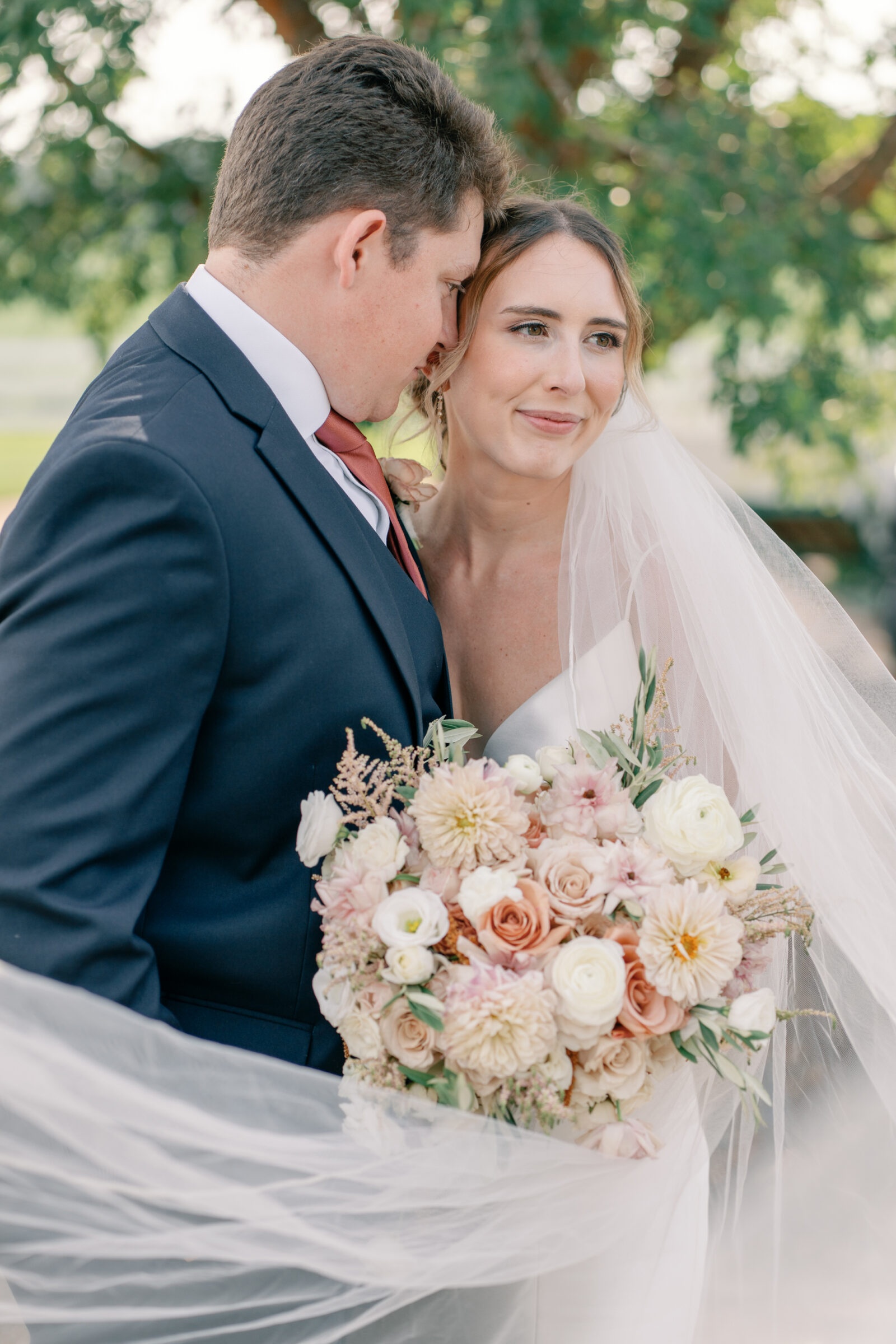 Bride with a flowing veil holding a pastel bouquet of dahlias and roses while the groom embraces her during wedding portraits at Burlington Weddings & Events in Charles City VA.
