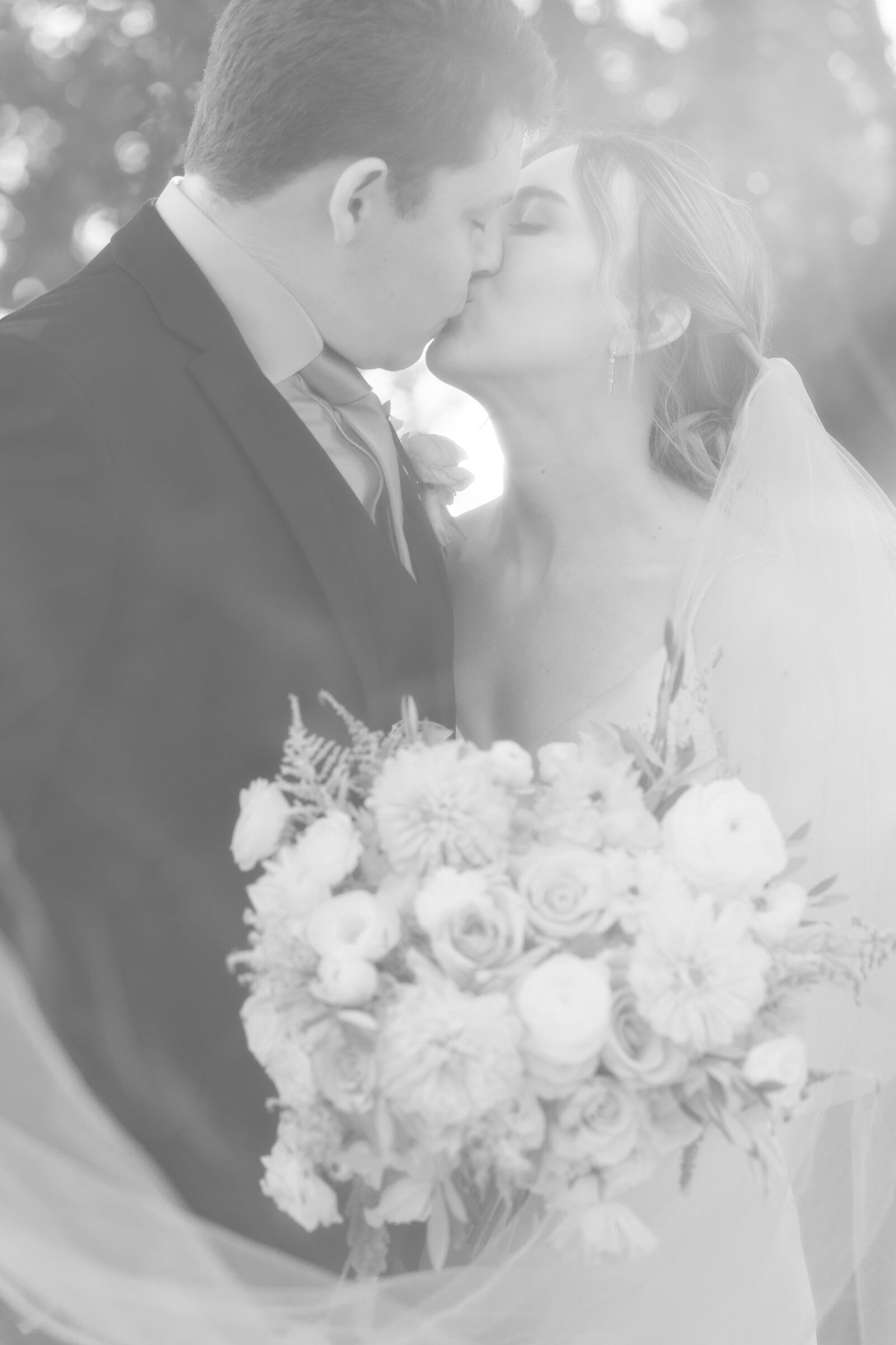 Black and white wedding photo of bride and groom sharing a kiss with the bride’s bouquet of roses and ranunculus.
