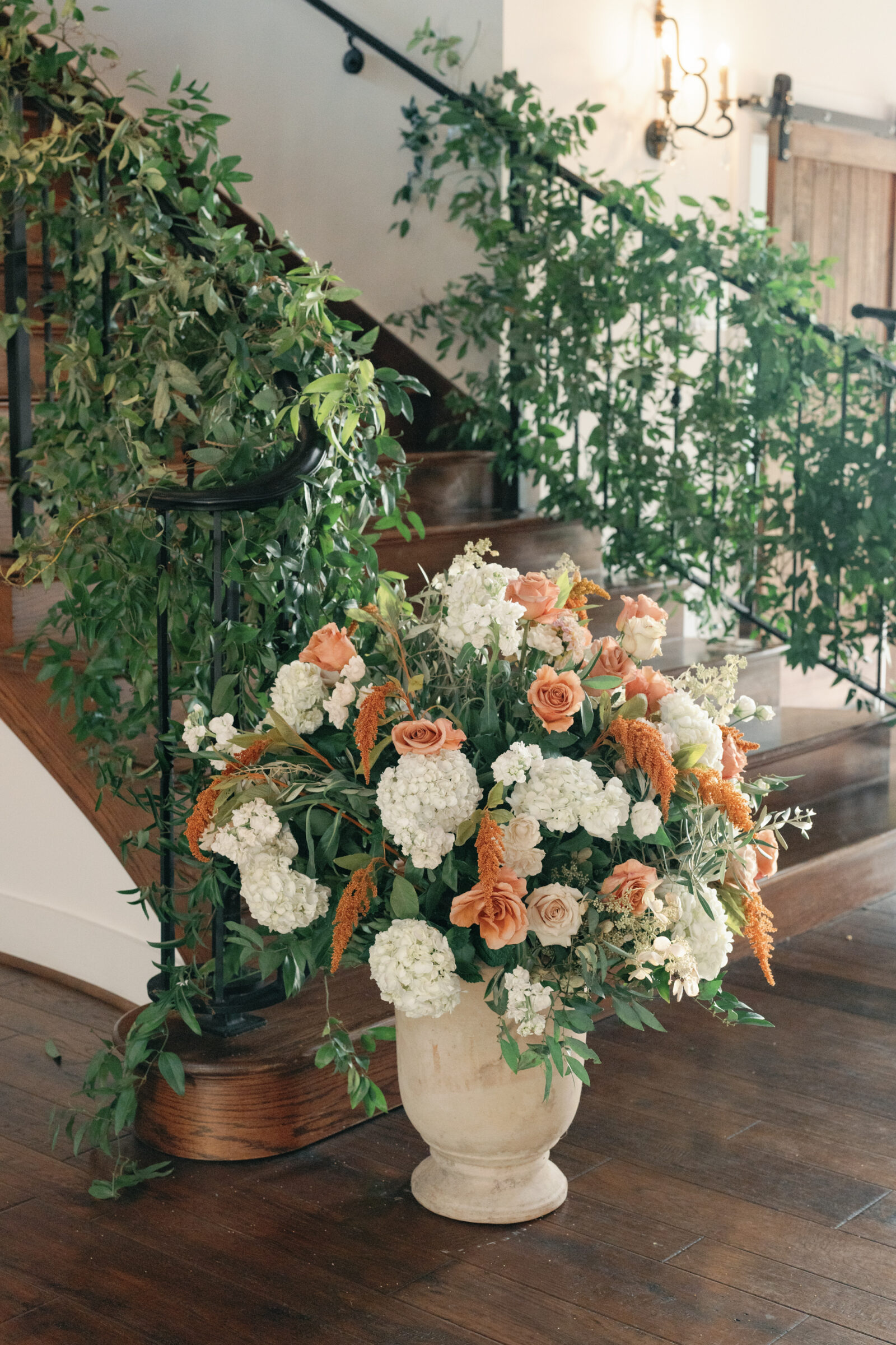 Large urn arrangement with white hydrangeas and peach roses beside a greenery-wrapped staircase.