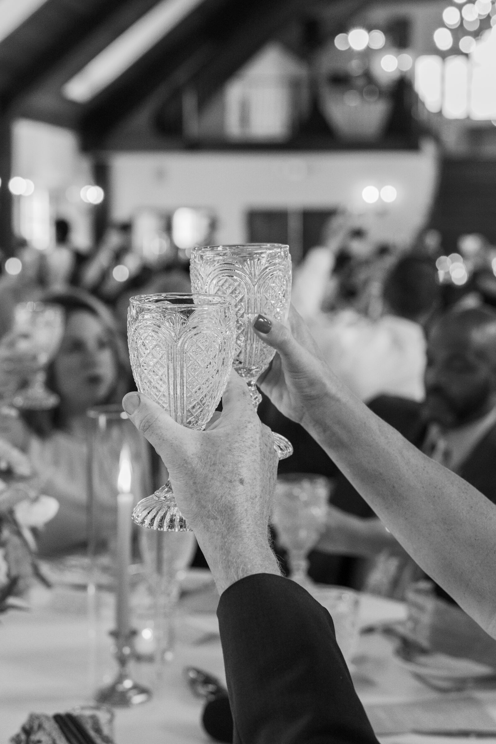 Black and white detail photo of guests raising vintage crystal glasses in a celebratory toast during a wedding reception.