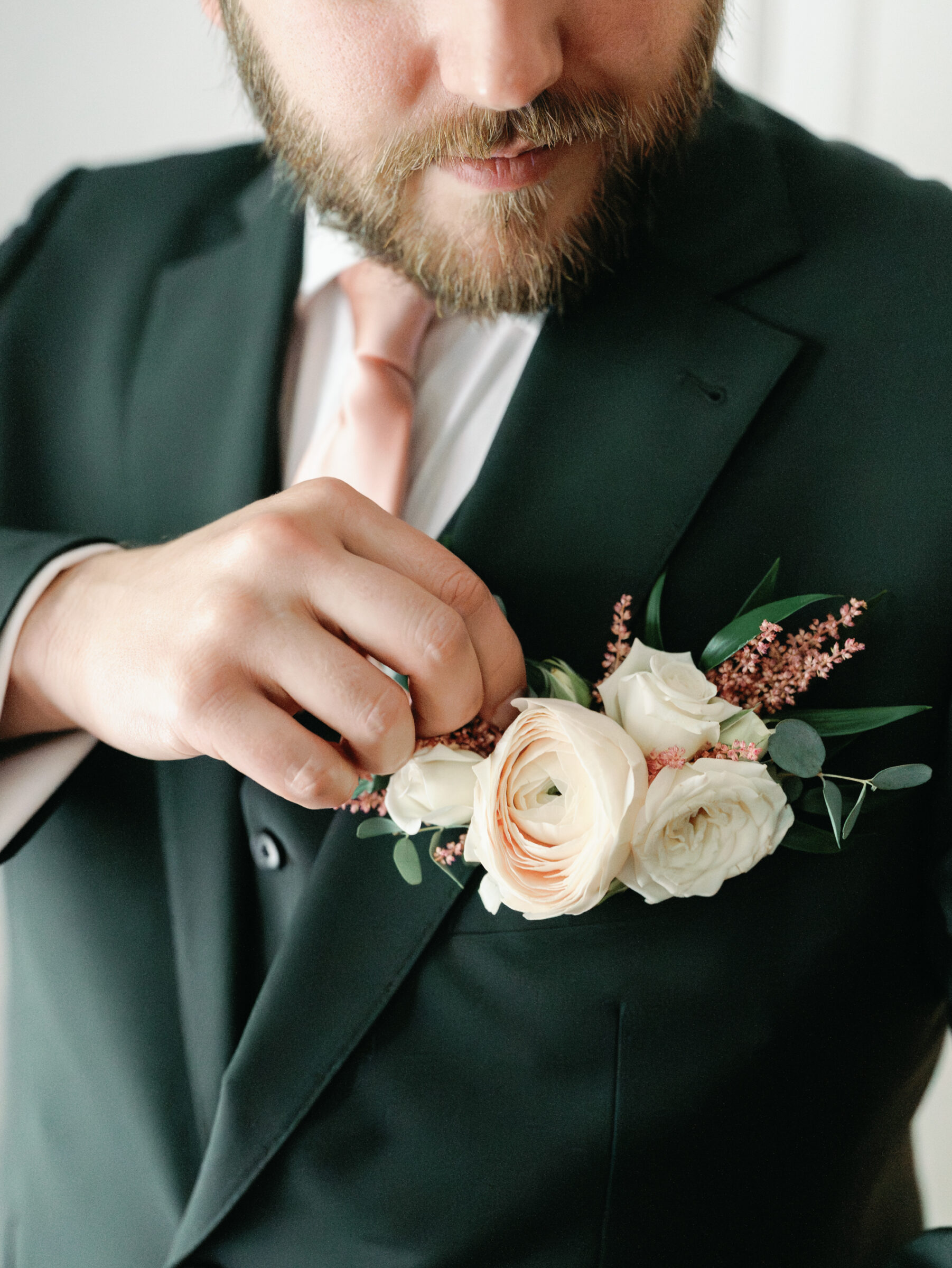 Groom in a forest green suit adjusting blush and white boutonniere with greenery before the ceremony.