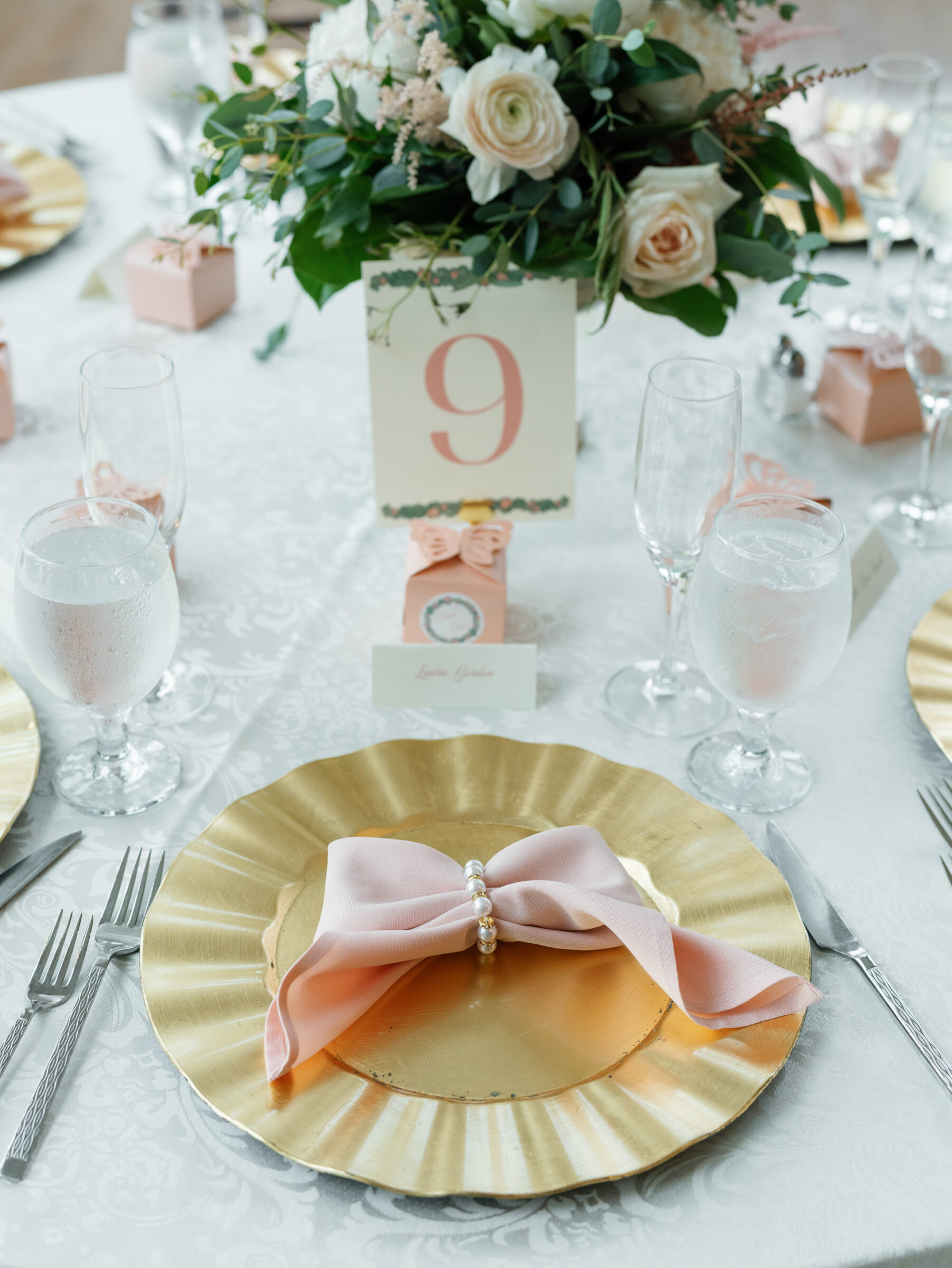 Elegant wedding reception table setting at Poplar Springs Manor with gold chargers, pink napkins tied with pearls, floral centerpiece, and table number card.