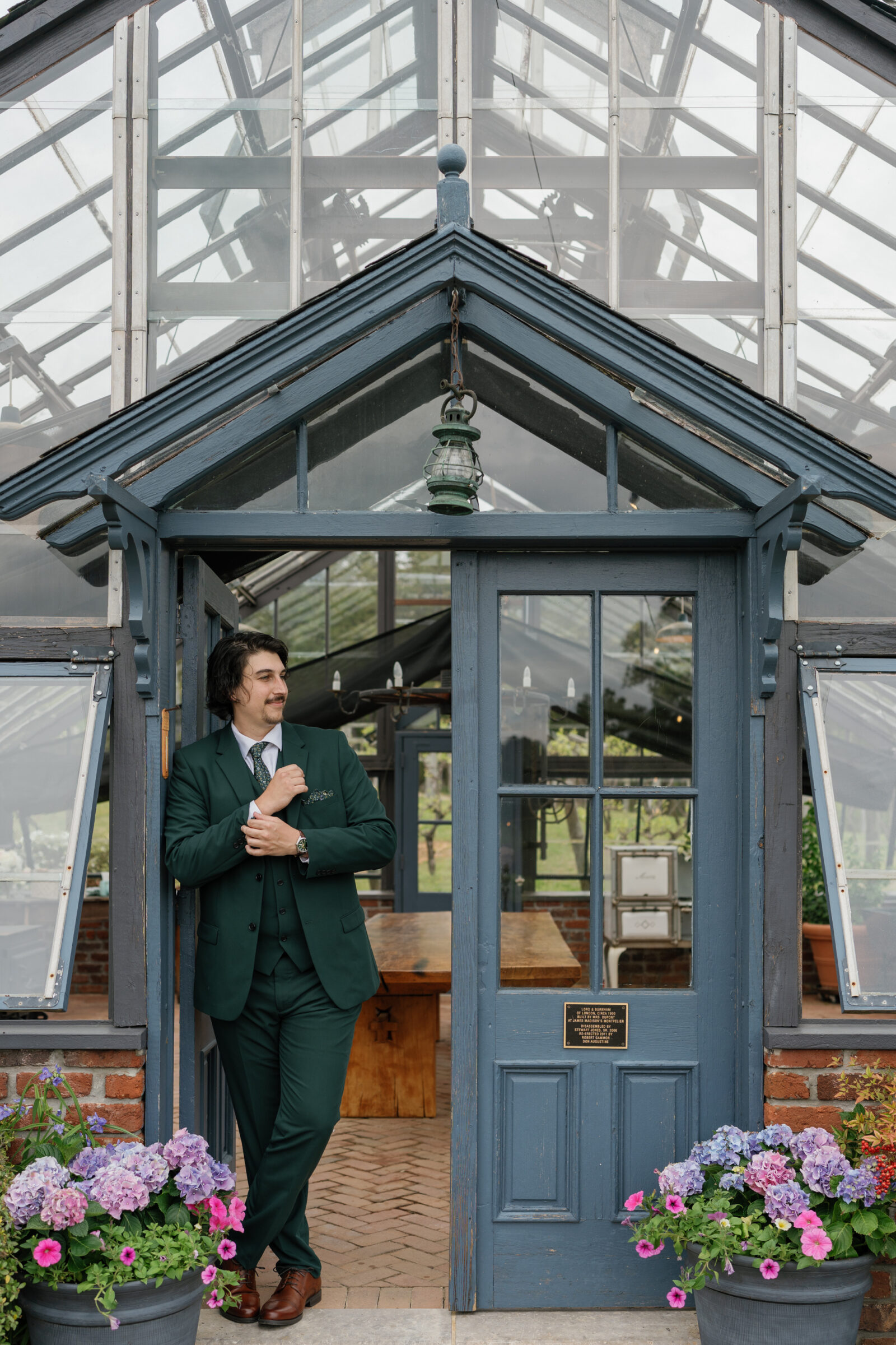 Groom in a dark green suit stands outside the greenhouse at Rassawek Vineyards in Columbia, Virginia, leaning casually against the blue doorway surrounded by vibrant hydrangeas, captured before his vineyard wedding.