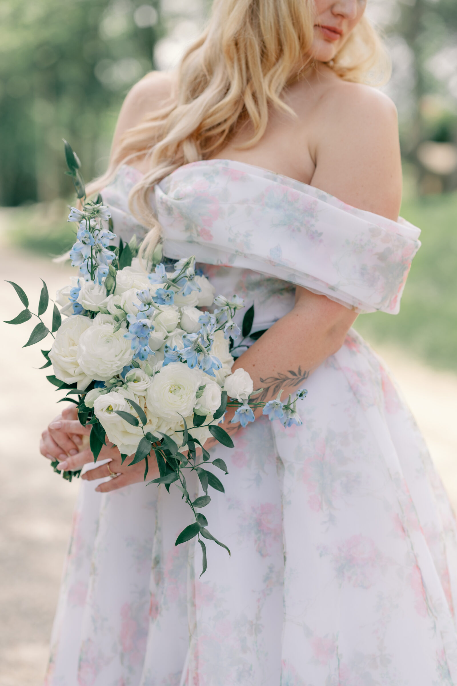 Bride in an off-the-shoulder floral wedding gown holds a romantic bouquet of white ranunculus and blue delphinium during her vineyard wedding in Virginia.