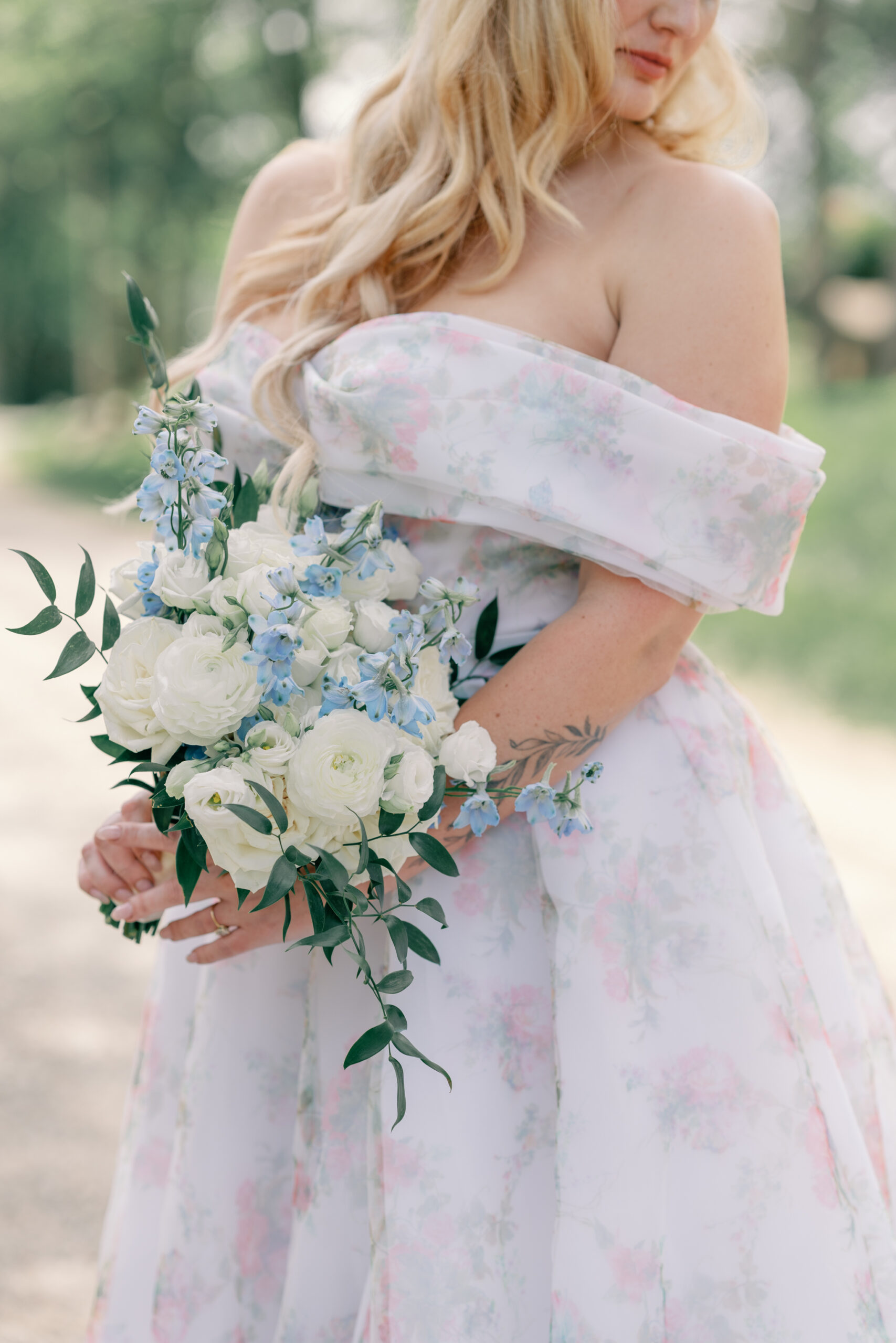 Bride in an off-the-shoulder floral wedding gown holds a romantic bouquet of white ranunculus and blue delphinium during her vineyard wedding at Rassawek Vineyards in Columbia, Virginia.
