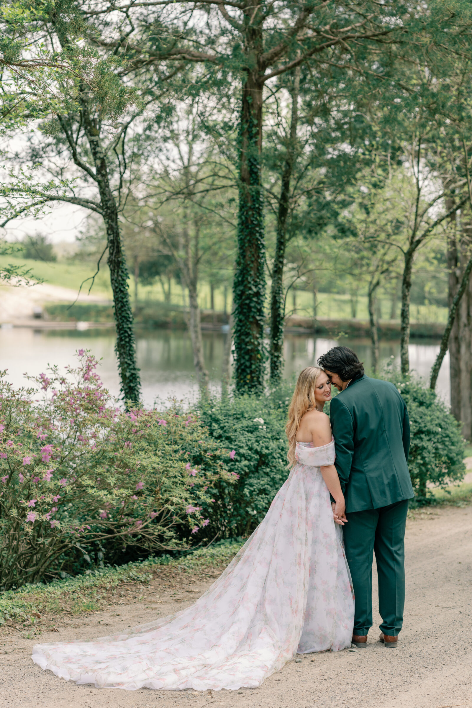 Bride in a floral off-the-shoulder gown walks hand in hand with the groom along the lakefront at Rassawek Vineyards in Virginia, her flowing train trailing behind during their vineyard wedding portraits.