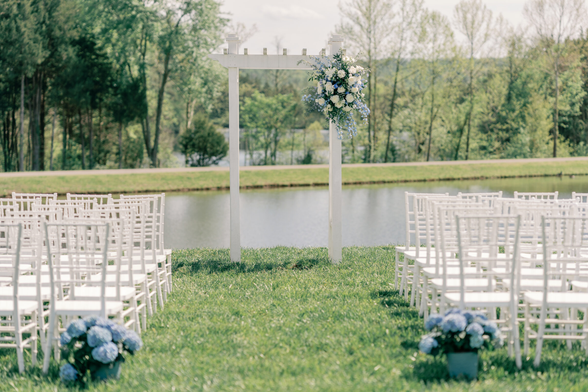 Outdoor wedding ceremony setup at Rassawek Vineyards in Virginia, featuring a white arbor with floral arrangements and rows of white chairs overlooking the lake.