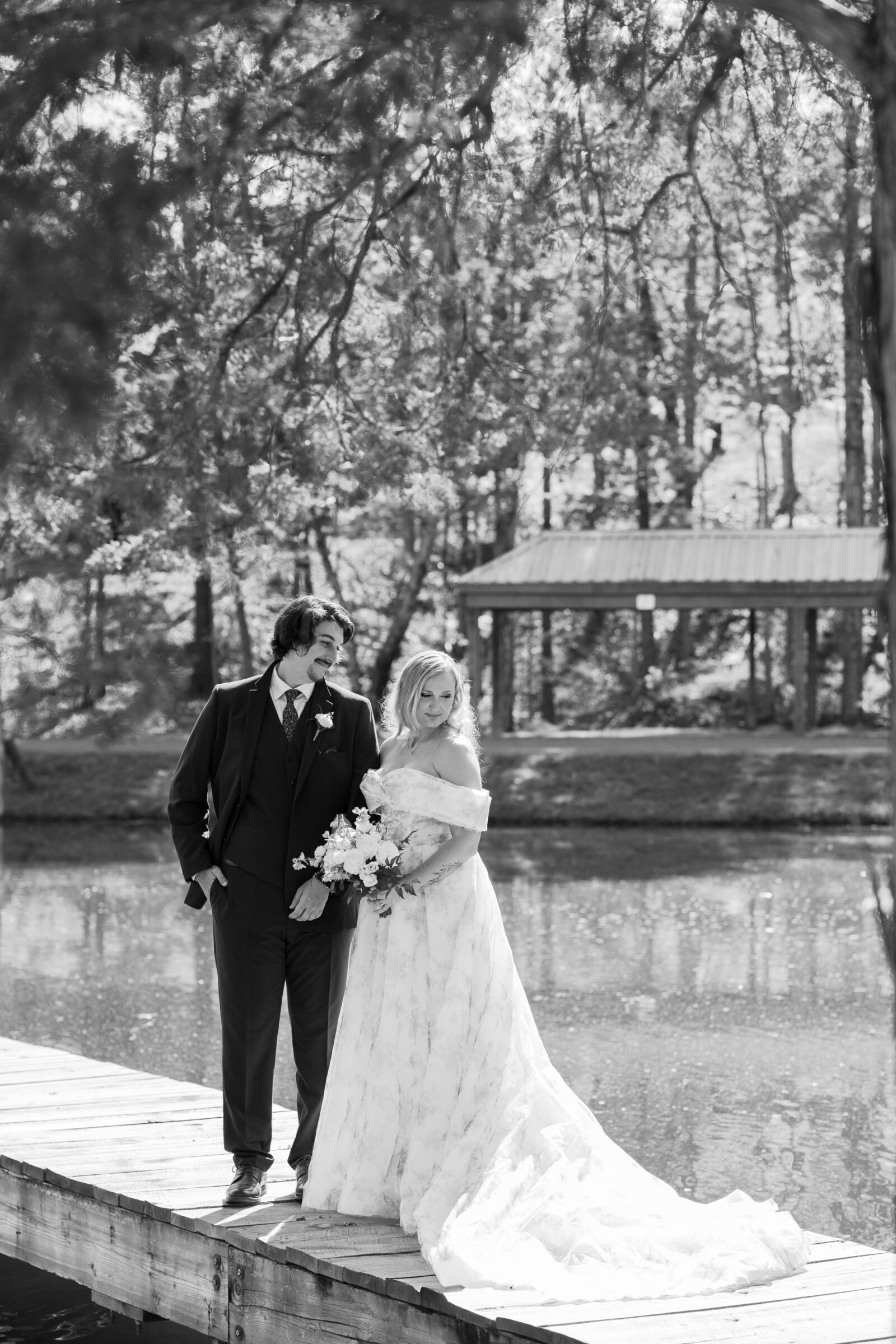 Bride and groom stand together on a wooden dock at Rassawek Vineyards in Virginia, captured in black and white with the bride holding a bouquet and her gown flowing beside the water.