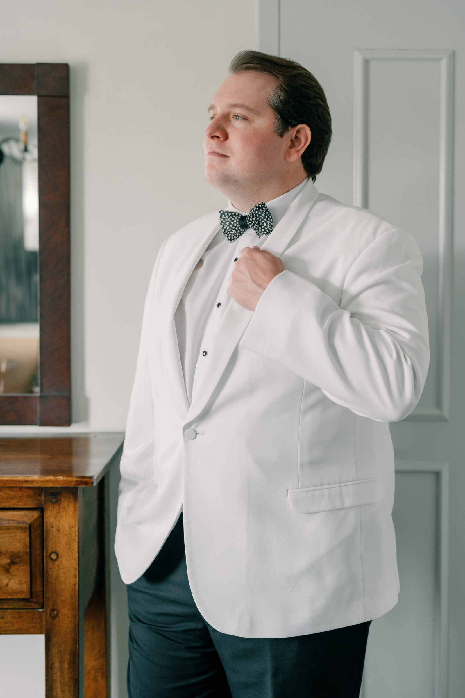 Groom getting ready for his Williamsburg wedding, wearing a white tuxedo jacket and black bow tie, adjusting his lapel indoors before the ceremony.