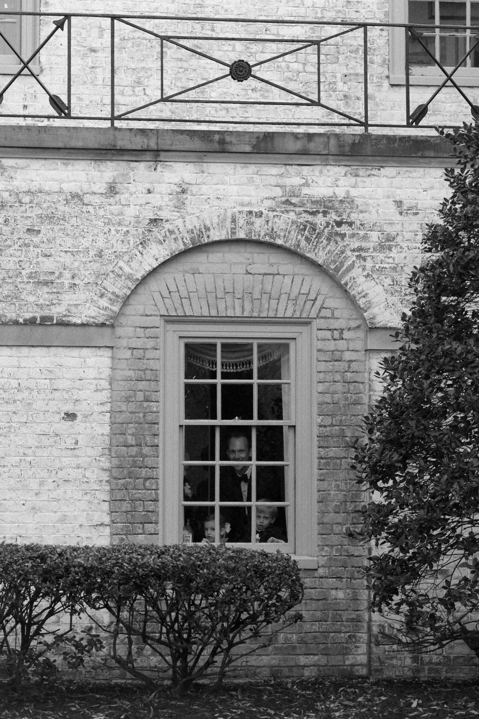 Candid black and white moment during a wedding with guests peering out the historic brick window, including a man in a tuxedo and two children.