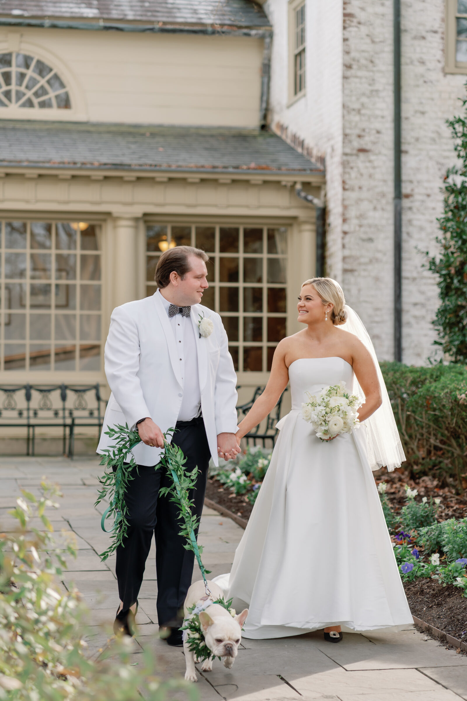 Bride and groom walking hand in hand with their dog during a Williamsburg Inn wedding, the bride holding a white bouquet and the groom in a white tuxedo jacket.