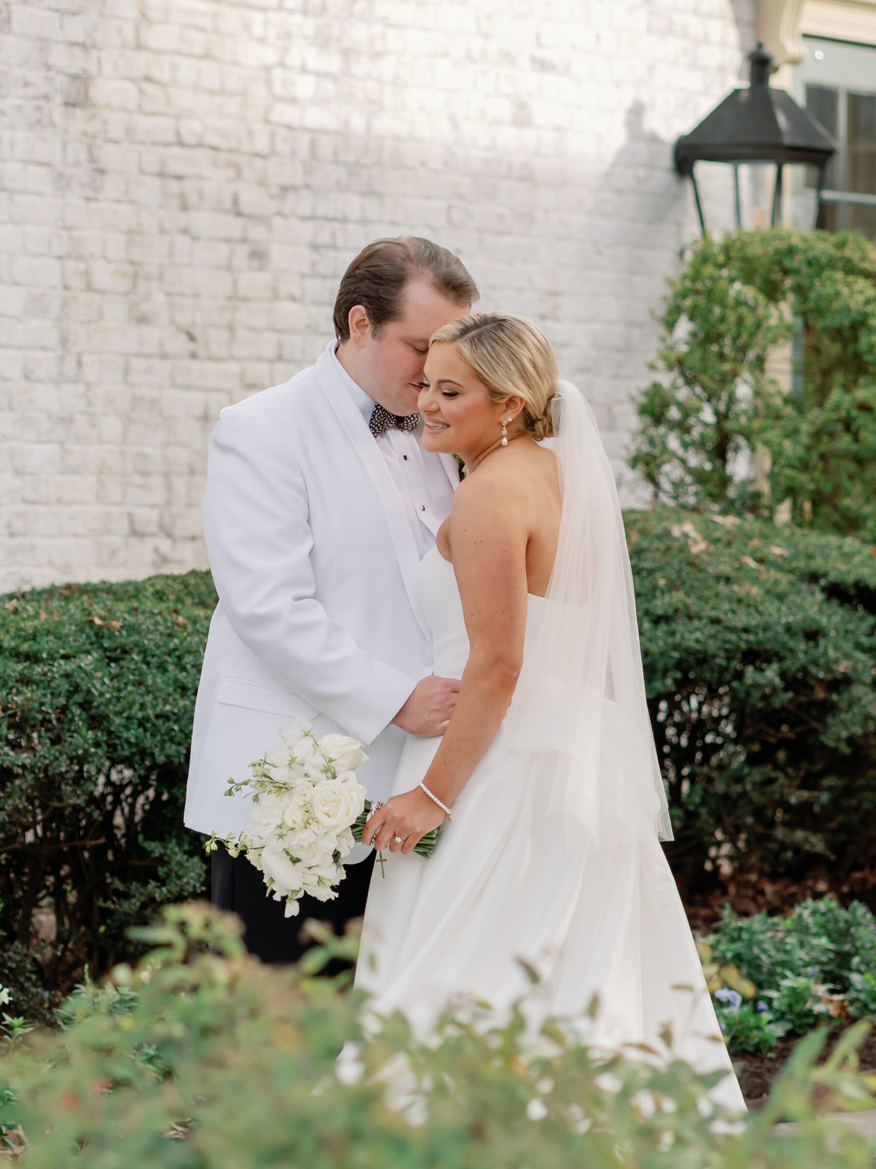 Bride and groom embracing during their Williamsburg Inn wedding, with the groom in a white tuxedo jacket and the bride smiling softly while holding a white bouquet.