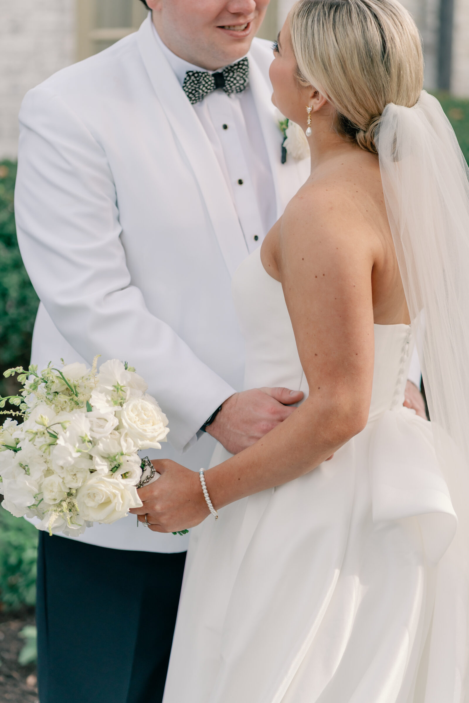 Close-up of the bride and groom during their Williamsburg Inn wedding, the bride holding a white rose bouquet and the groom in a white tuxedo jacket with a patterned bow tie.