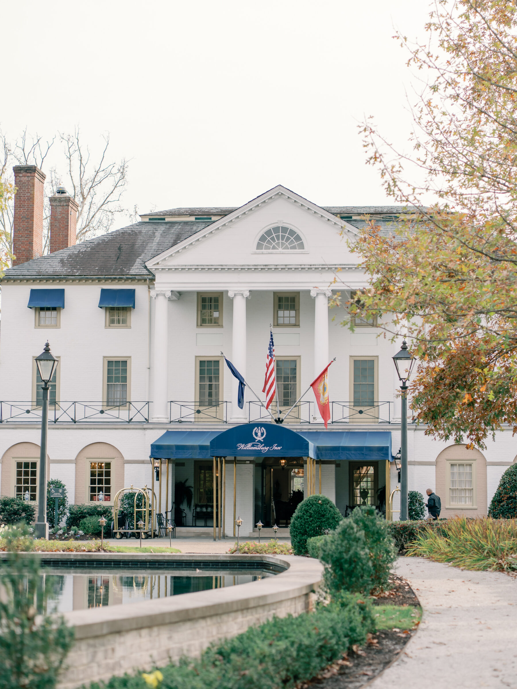 Exterior view of the Williamsburg Inn wedding venue, featuring its white brick façade, grand columns, blue awnings, and reflecting pool at the entrance.
