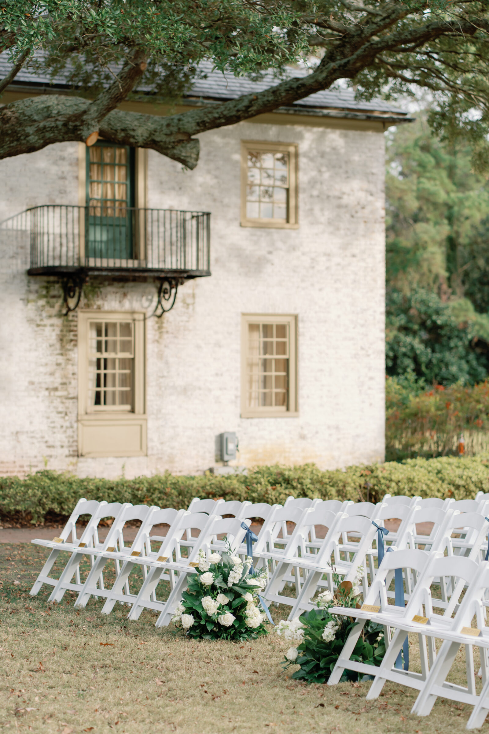 Outdoor ceremony setup at a Williamsburg Inn wedding with rows of white chairs and floral arrangements of white roses and greenery under a large oak tree.