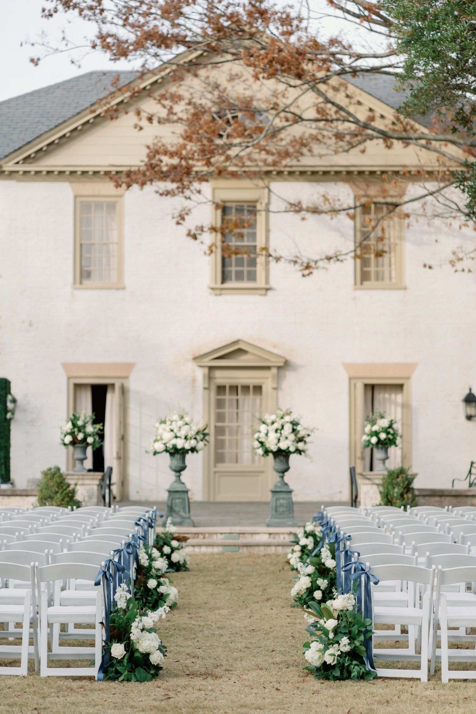 Elegant outdoor ceremony space at a Williamsburg Inn wedding with rows of white chairs, navy ribbon accents, and large white floral arrangements framing the altar.
