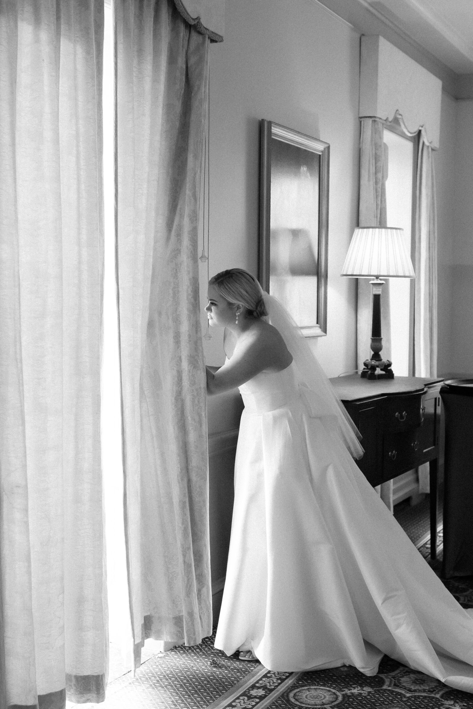 Black and white photo of a bride in her gown peeking through the curtains before her wedding ceremony.