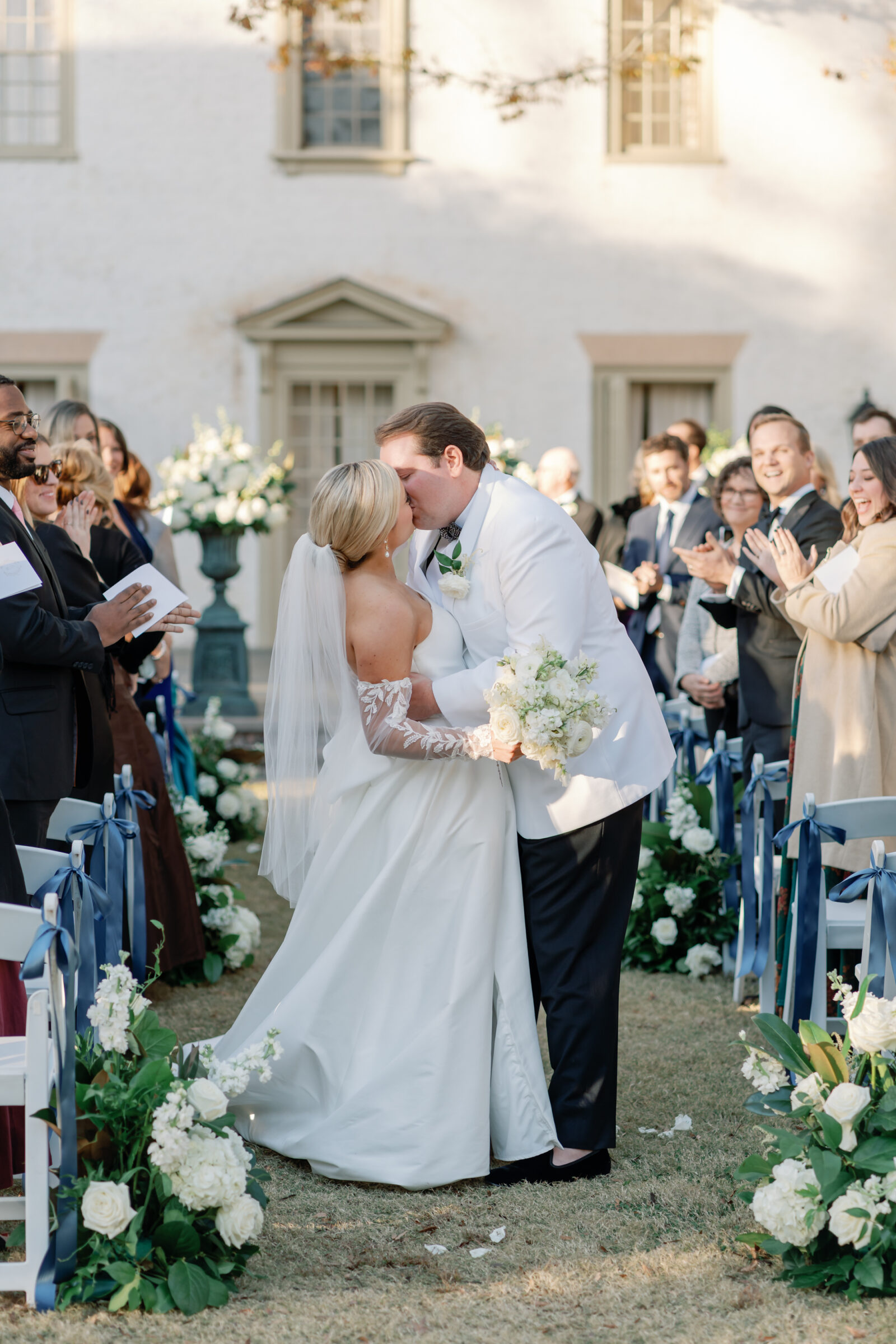 Bride and groom share their first kiss as husband and wife during an outdoor Williamsburg Inn wedding ceremony, surrounded by guests applauding and floral arrangements.