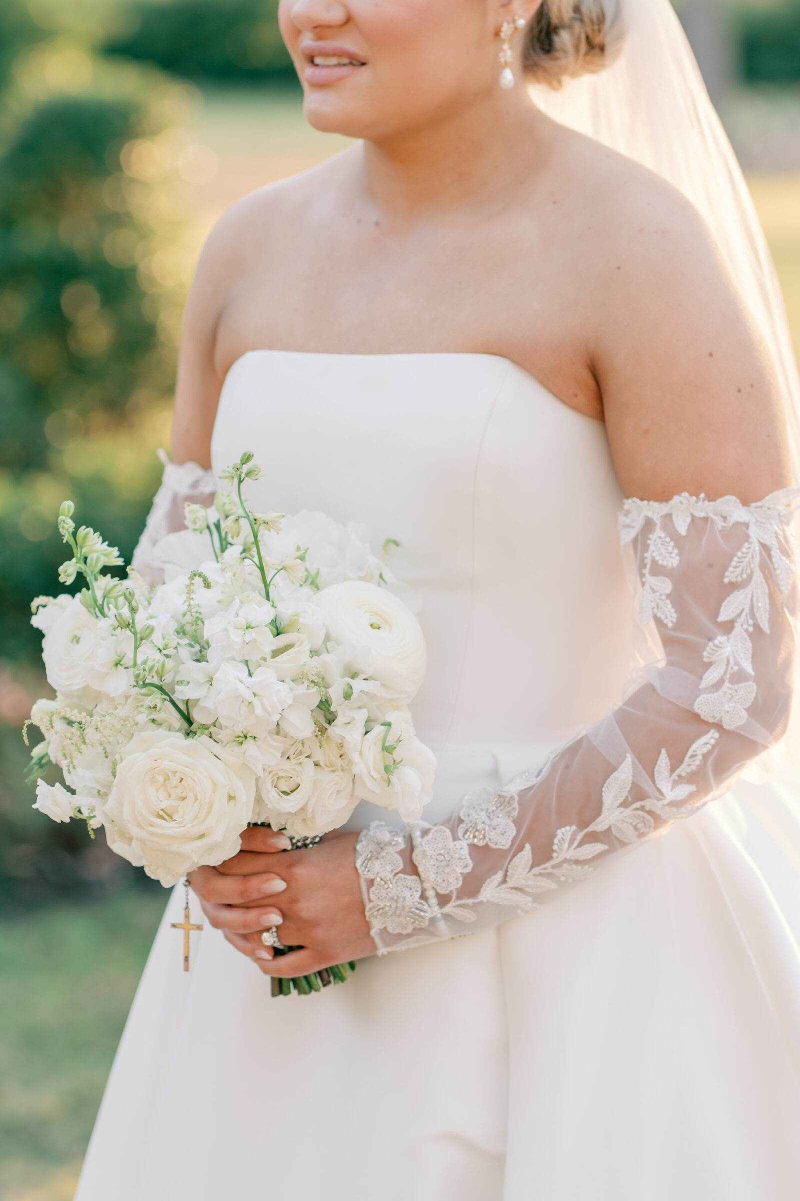 Close-up of the bride holding a white floral bouquet, wearing a strapless gown with lace illusion sleeves and pearl earrings.