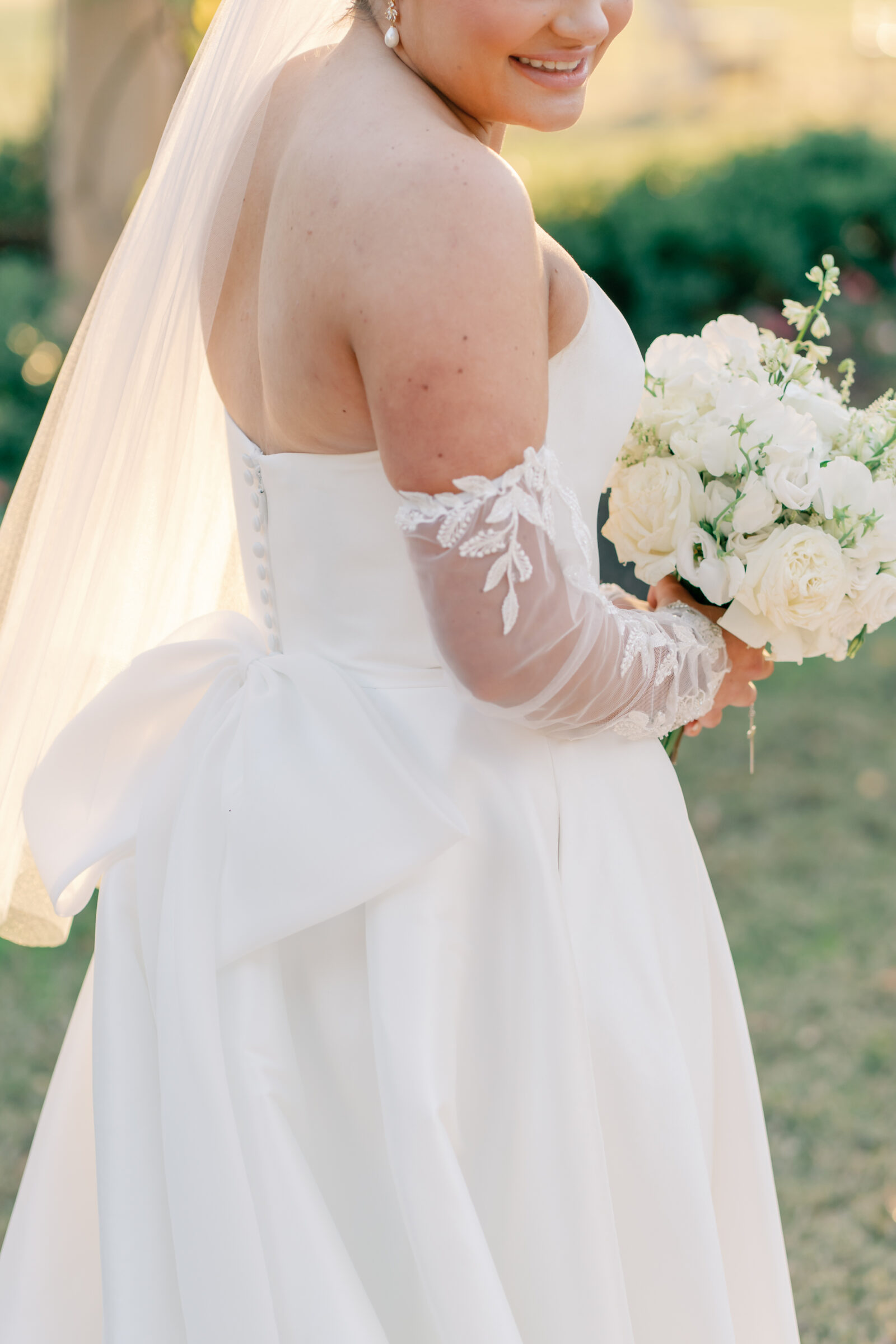 Bride holding a white rose bouquet, with sunlight highlighting the bow detail on the back of her gown and lace sleeves.
