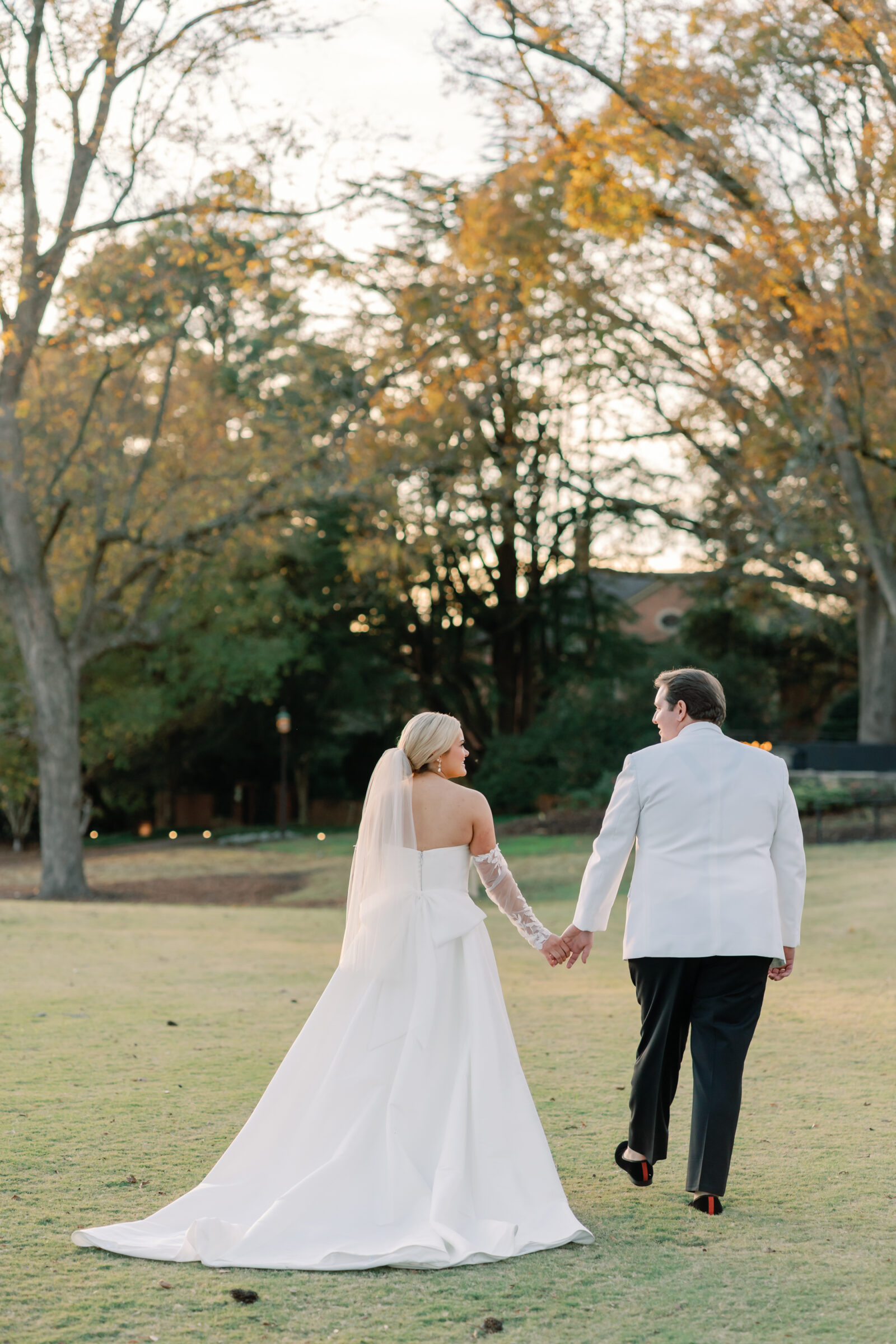 Bride and groom walking hand in hand across the lawn at their Williamsburg Inn wedding, with autumn trees glowing in the golden hour light.