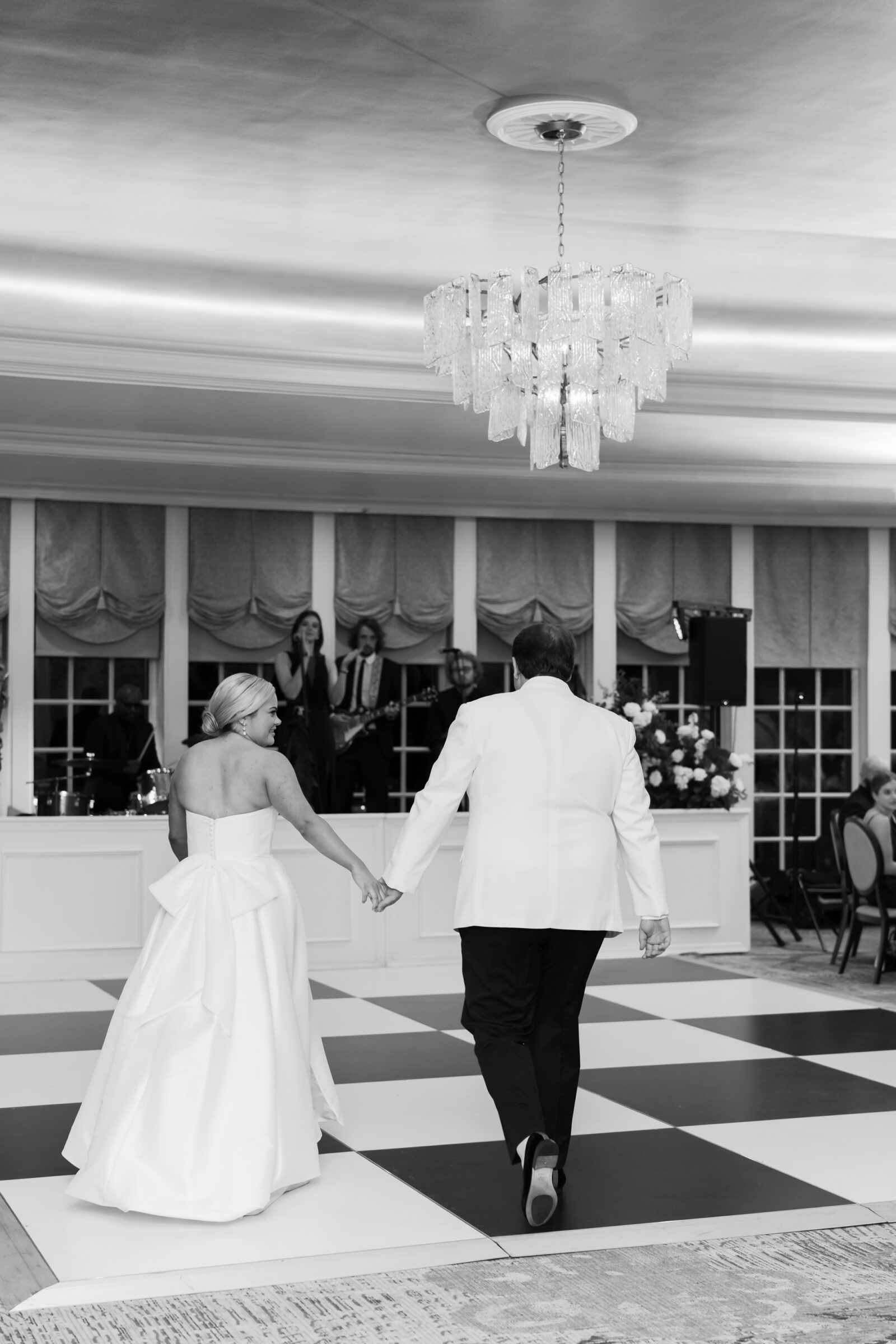 Bride and groom holding hands as they enter the reception ballroom for their first dance at their Williamsburg Inn wedding, with live band performing on stage.