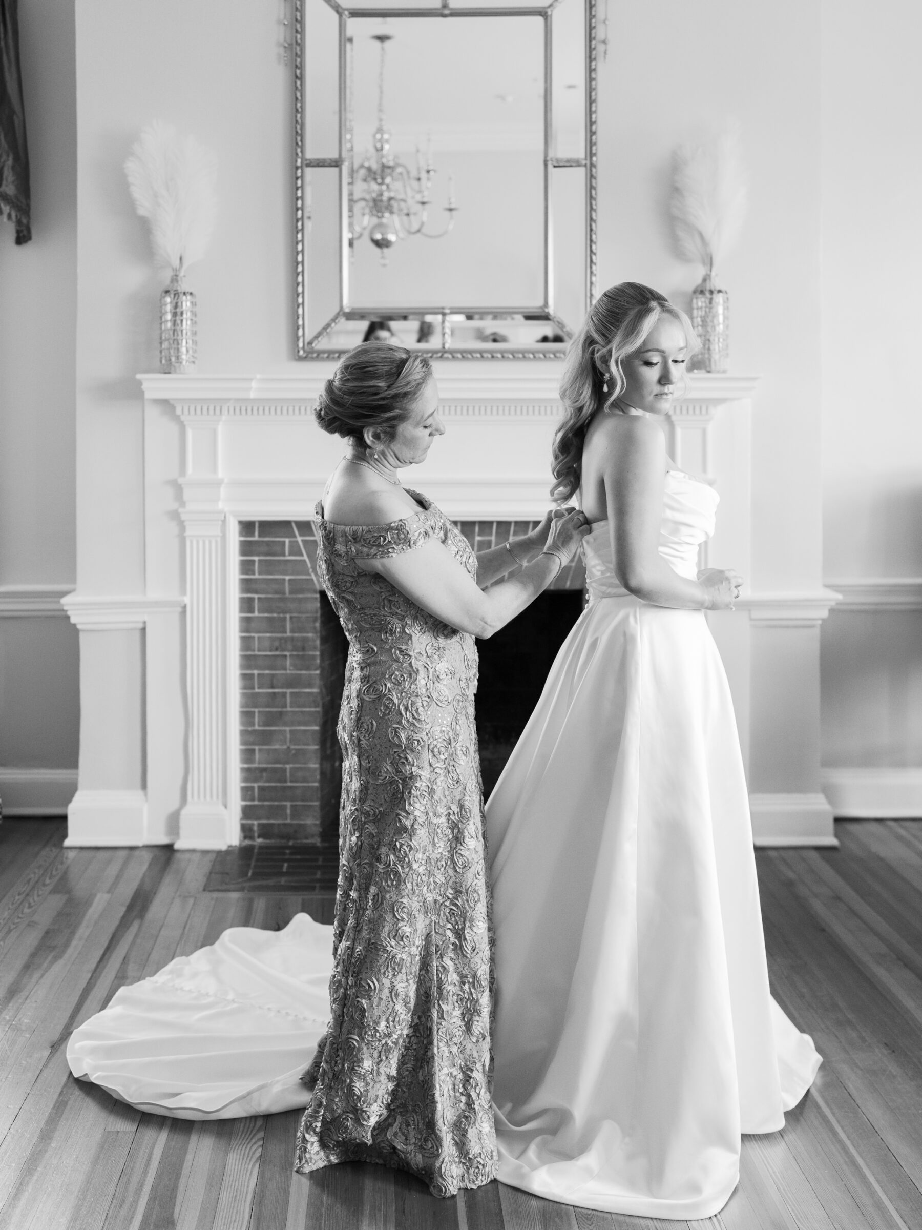 Bride standing by the fireplace as her mother fastens her wedding gown in the bridal suite at Belmont Country Club in Ashburn, VA.