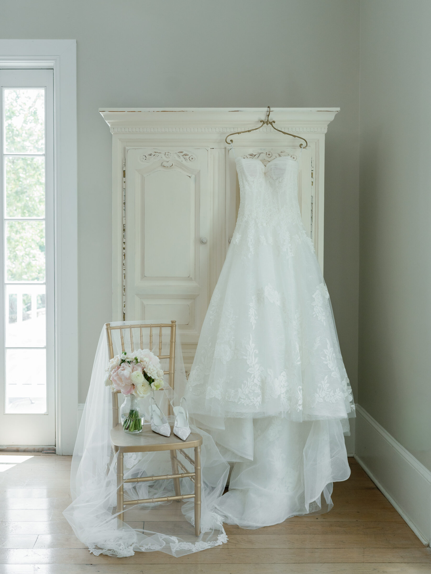A lace strapless wedding gown hangs on a vintage white armoire with the bride’s bouquet, veil, and shoes styled on a chair beside it in soft natural light.