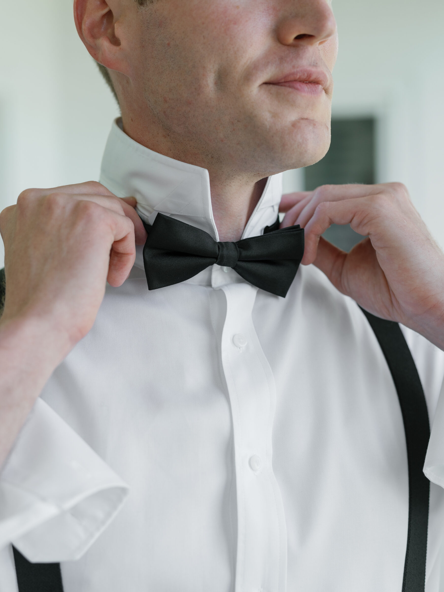 Close-up of the groom adjusting his black bow tie while wearing a white dress shirt and suspenders before the ceremony.