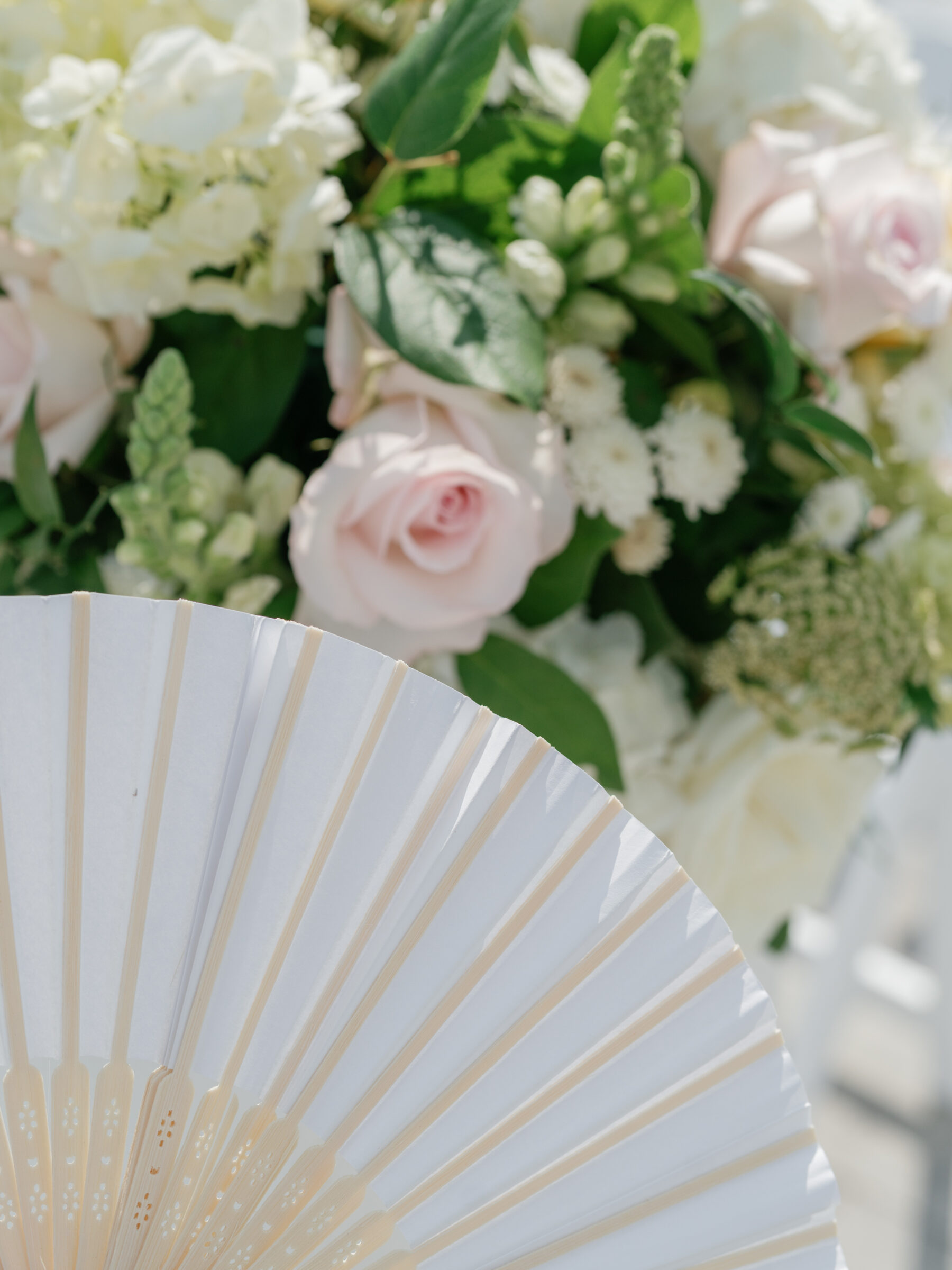 Close-up of pastel roses and white florals beside an elegant handheld fan for guests at a summer wedding ceremony.