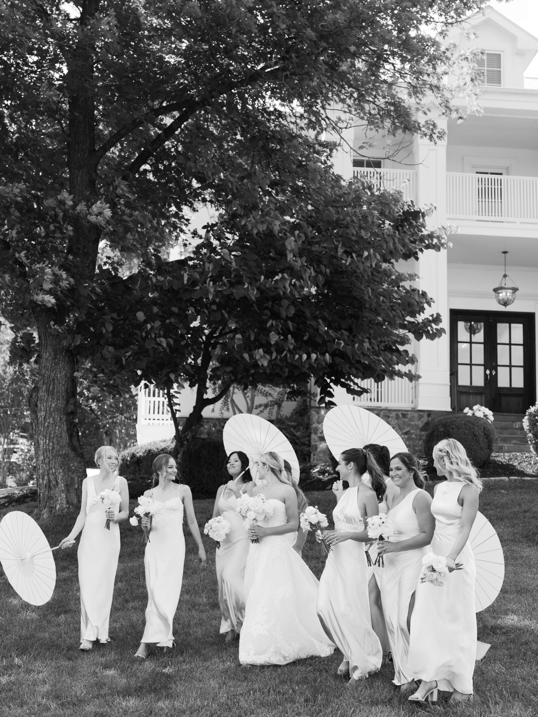 A bridal party in soft summer gowns walking with parasols on the lawn of the manor house at a timeless The View at Bluemont Wedding.