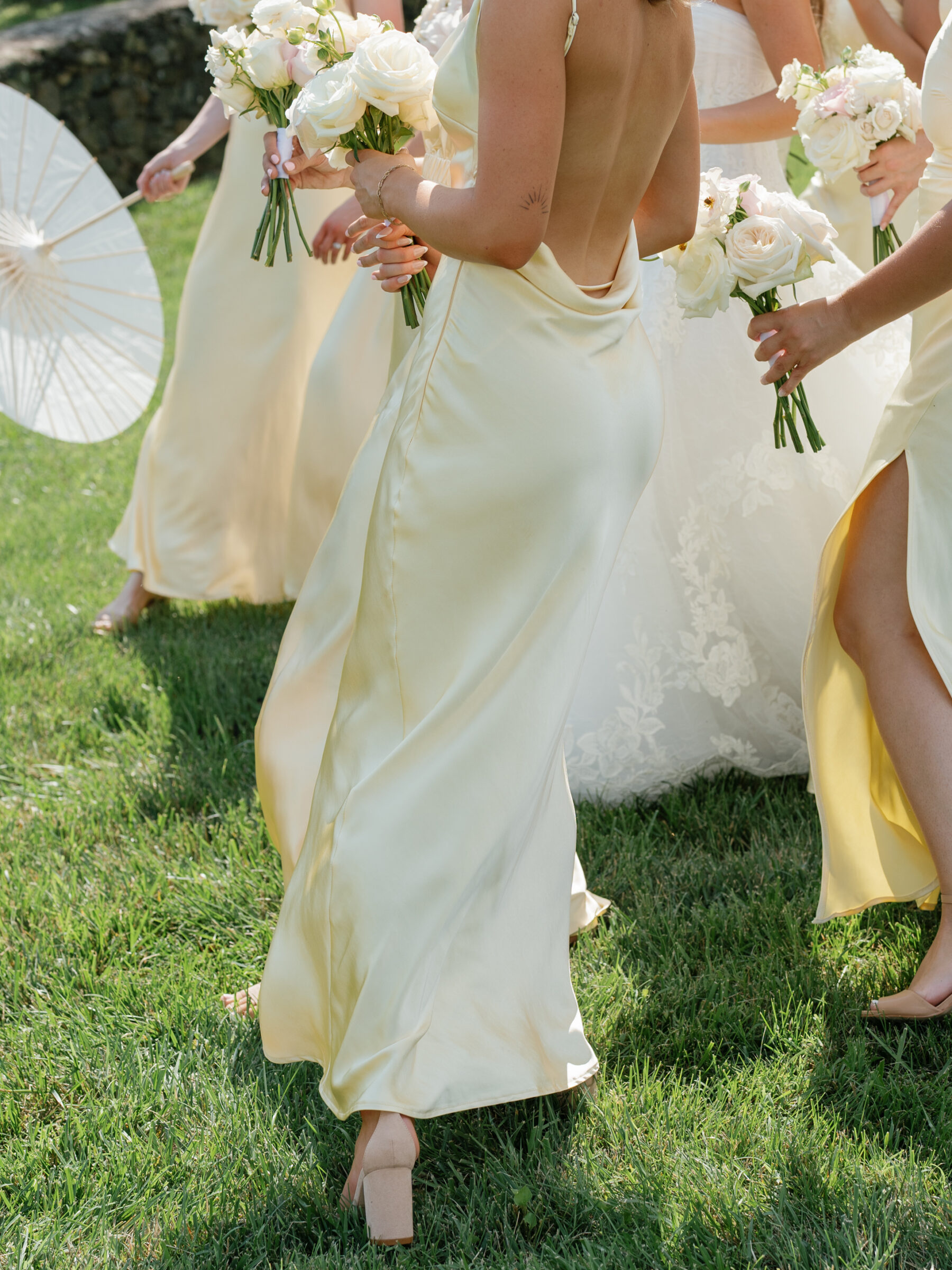 Bridesmaids in soft yellow satin gowns walking across the grass holding white rose bouquets next to the bride.