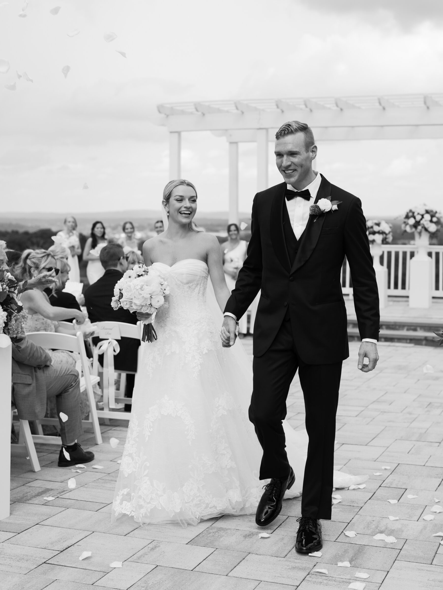 The newlyweds smiling and walking up the aisle together as guests toss flower petals during their outdoor ceremony celebration.