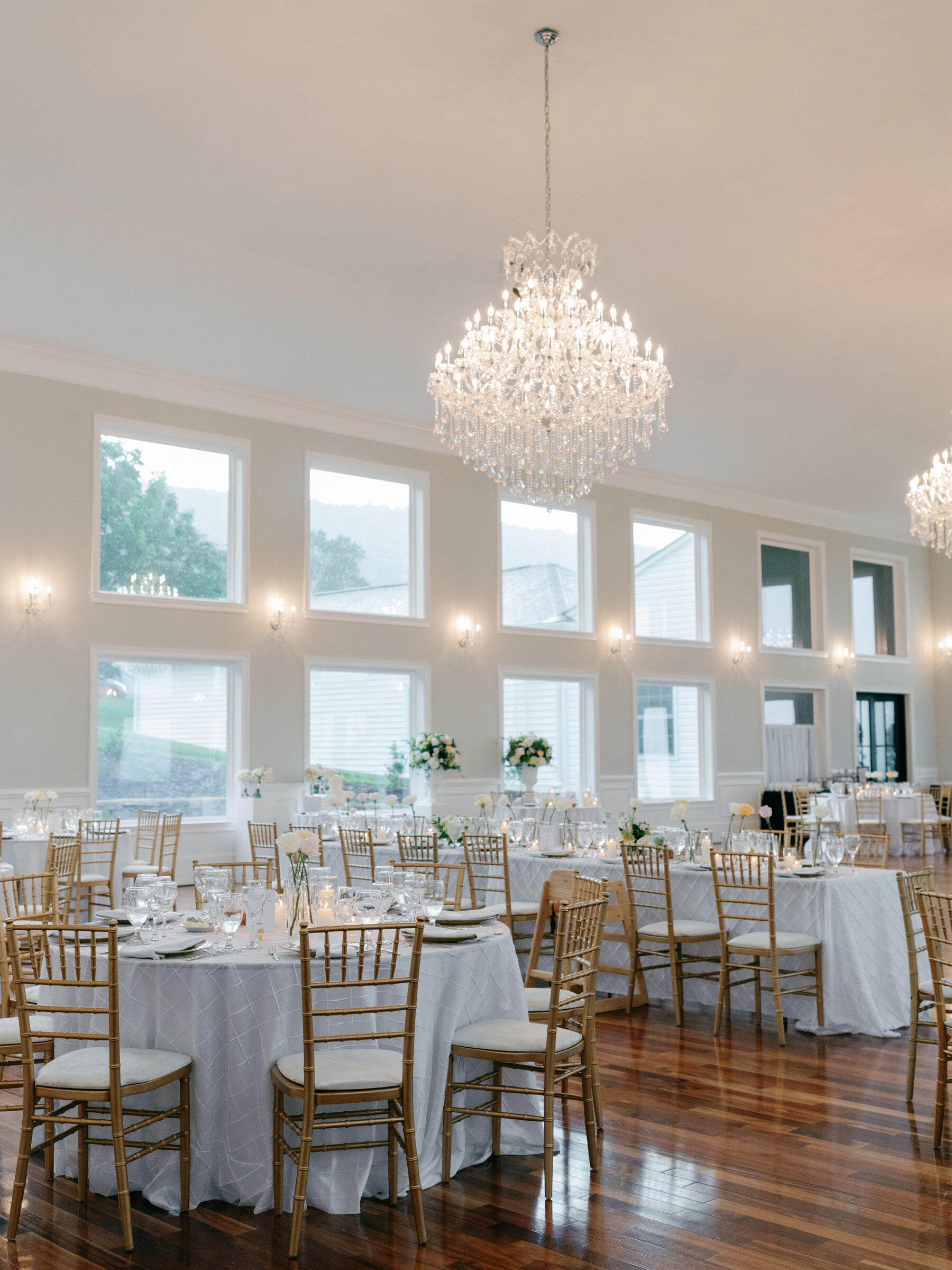 Wide view of an elegant wedding reception setup with gold chairs, candlelit tables, and crystal chandeliers in a bright ballroom.