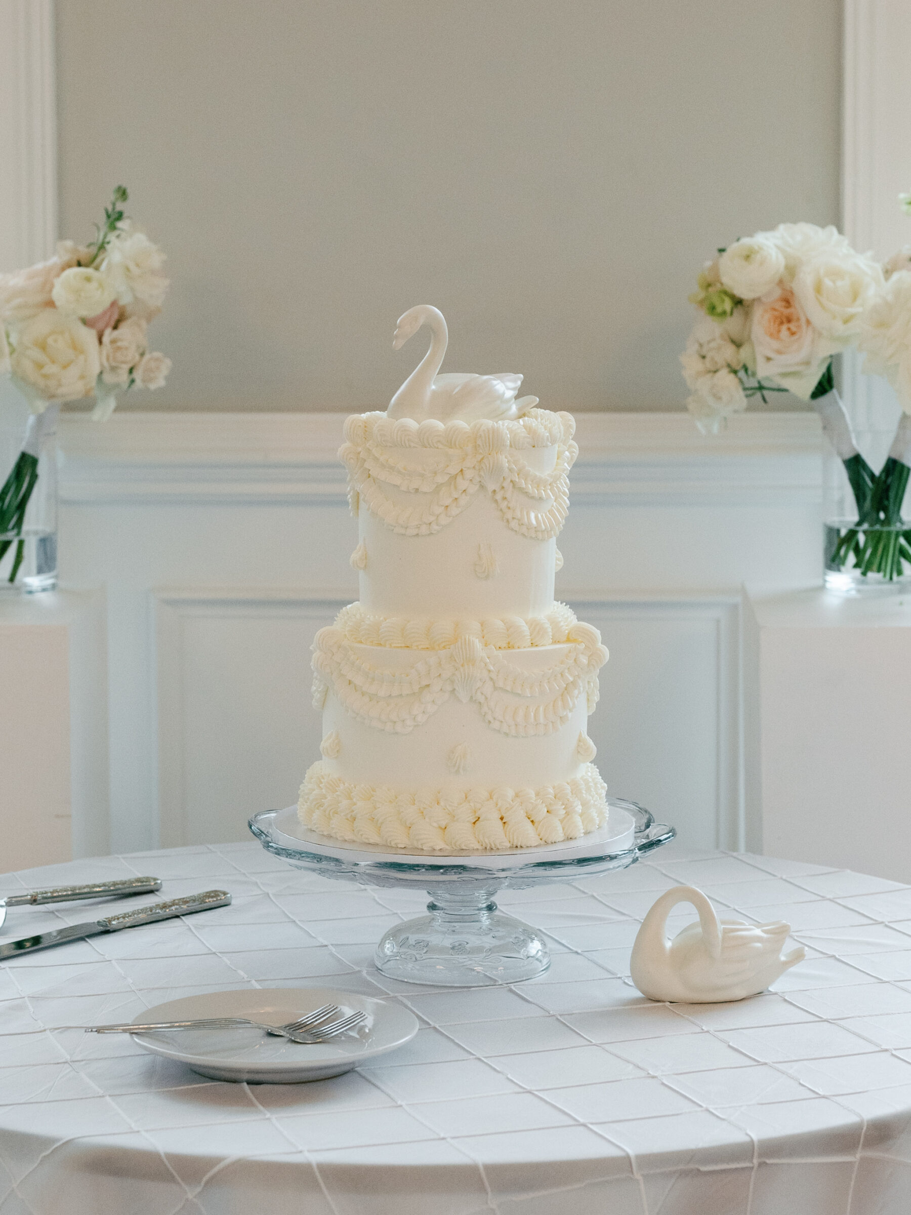 A classic white wedding cake with vintage-inspired piping and a porcelain swan topper displayed among romantic florals and candlelight.