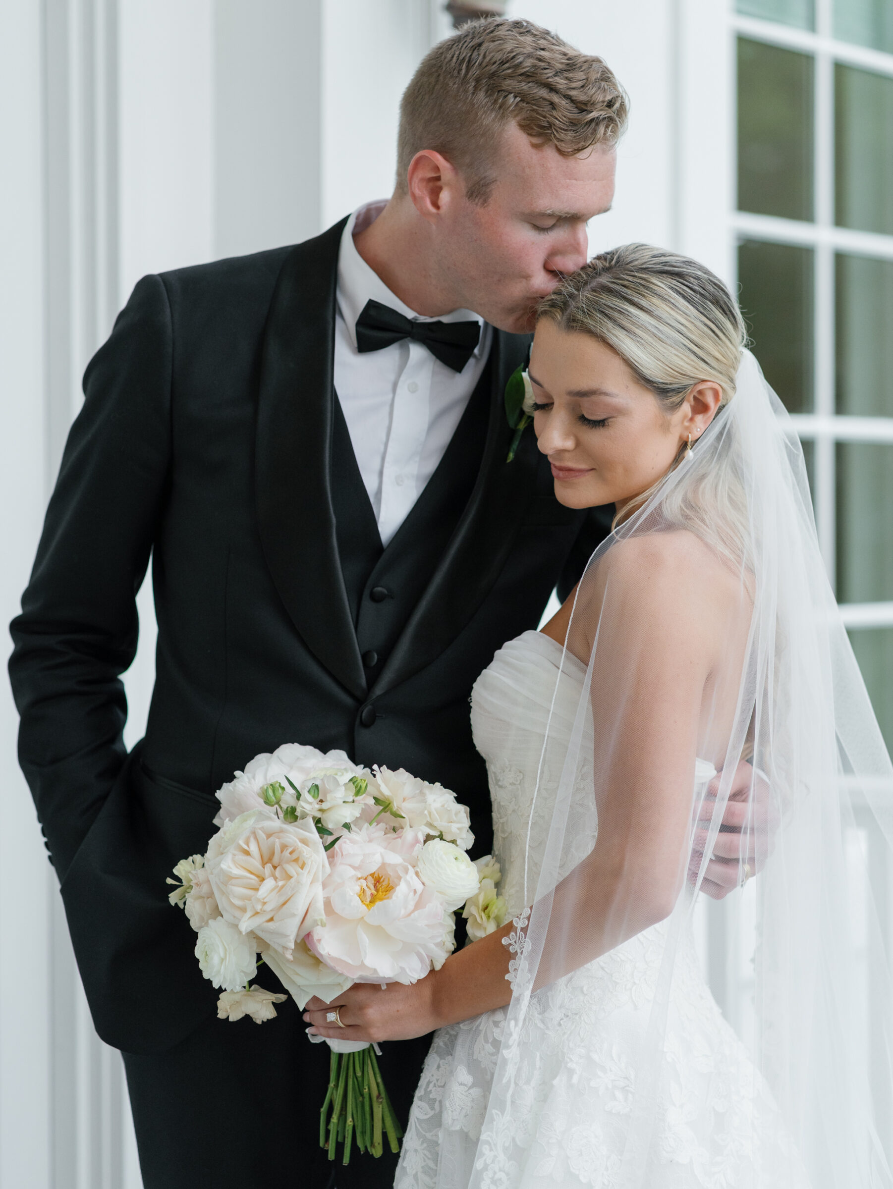 The groom gently kisses the bride on the forehead as she holds a soft pastel bouquet, both wrapped in an intimate embrace.