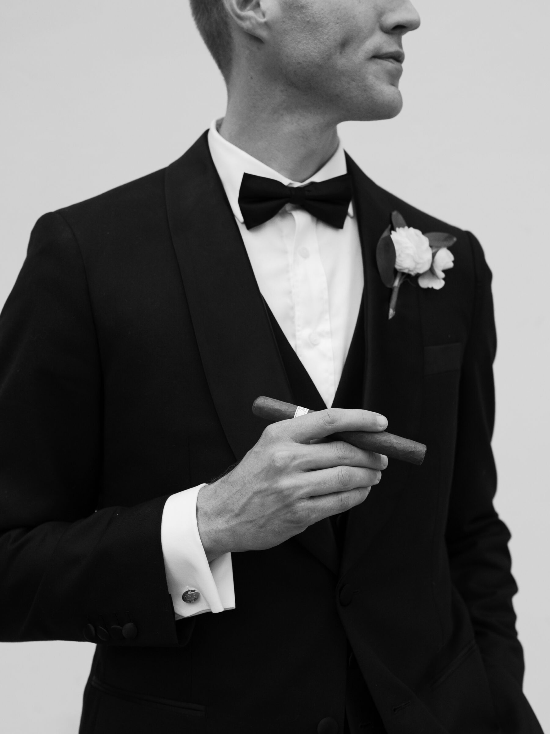 Stylish groom portrait with a cigar, black bow tie, and classic tuxedo details in black and white.