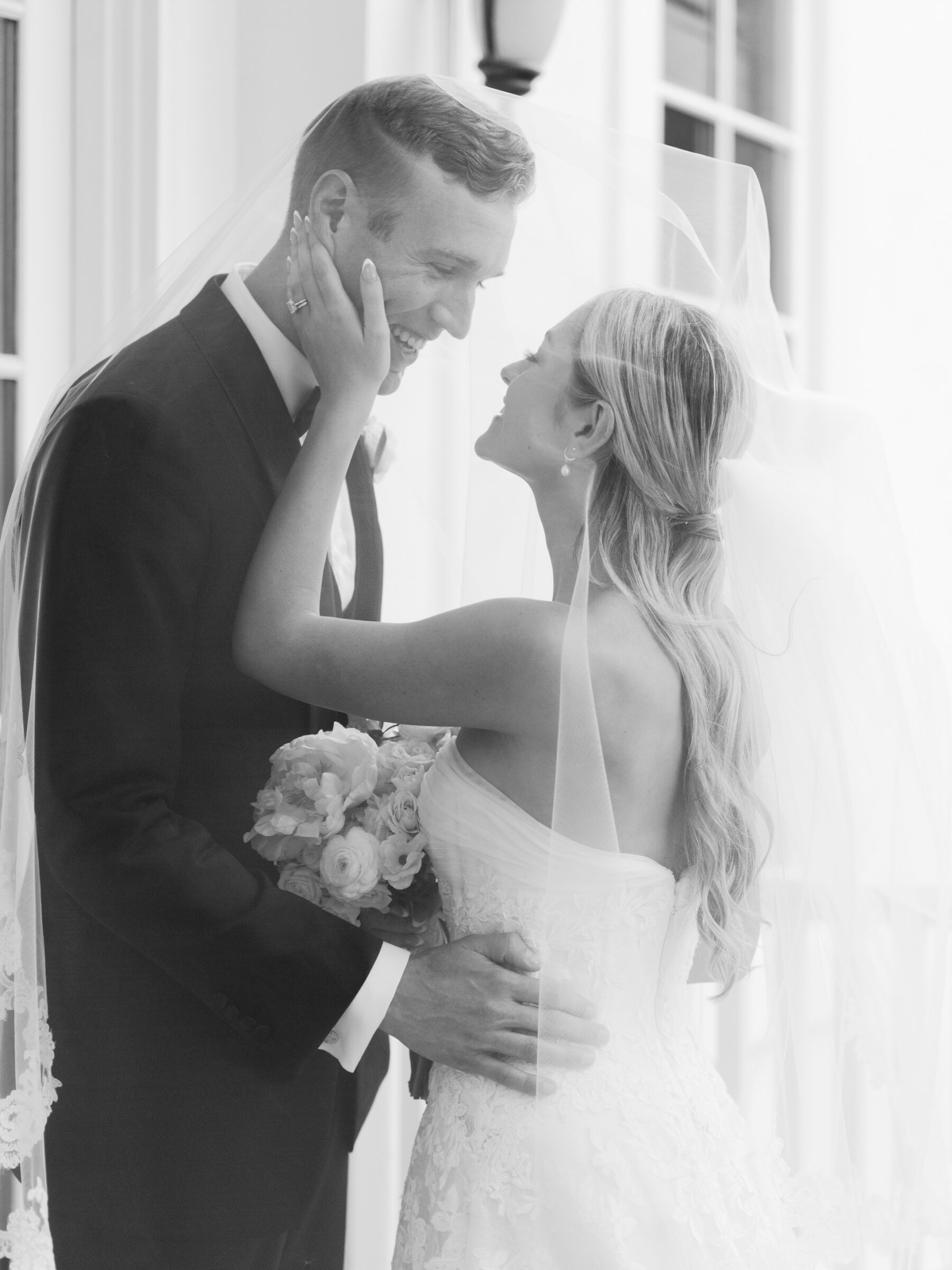 The bride gently holds the groom’s face as they laugh together beneath her veil, wrapped in an intimate embrace.