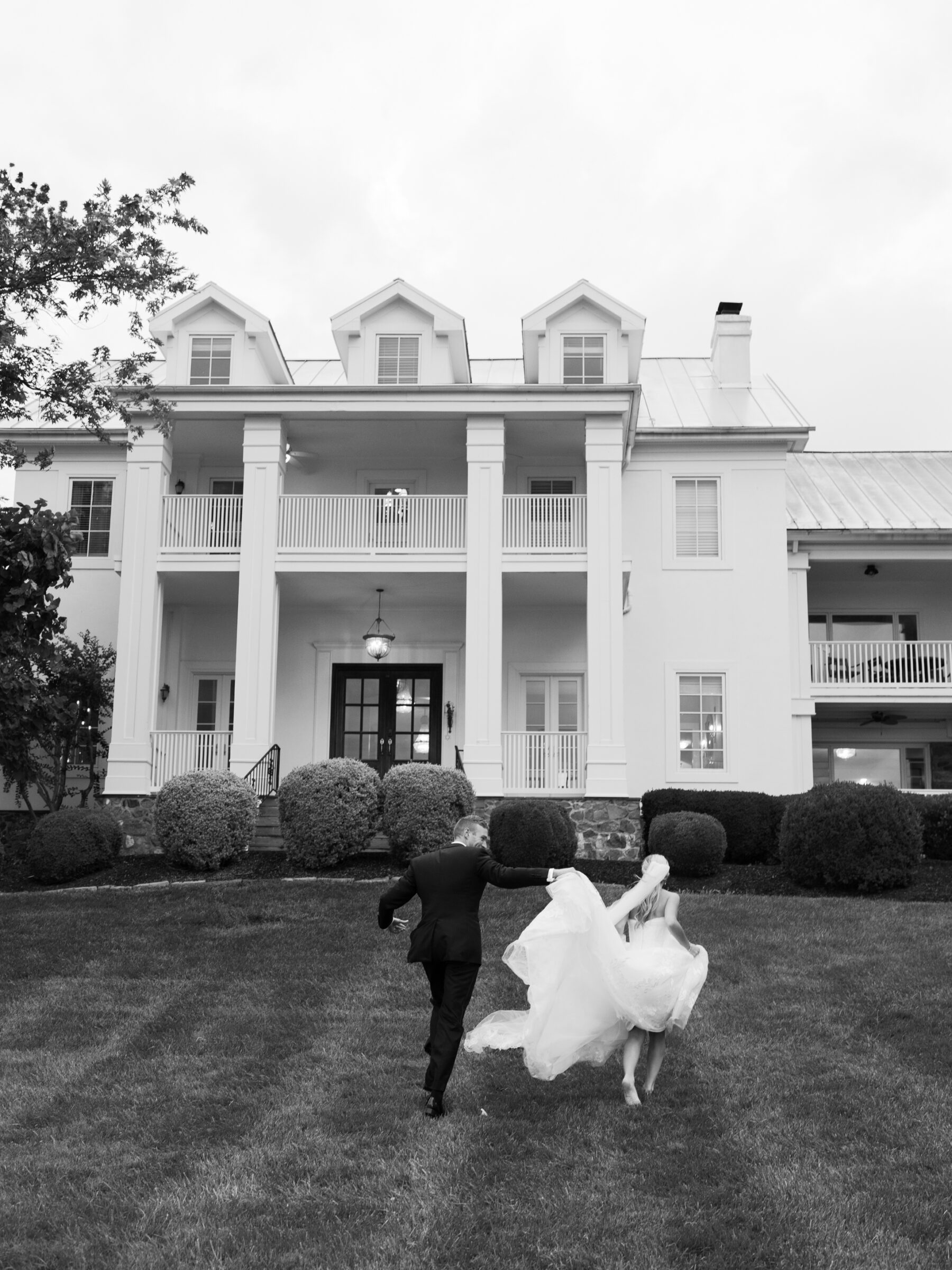 The bride and groom run hand-in-hand toward the grand white estate, her gown flowing behind them in joyful motion.