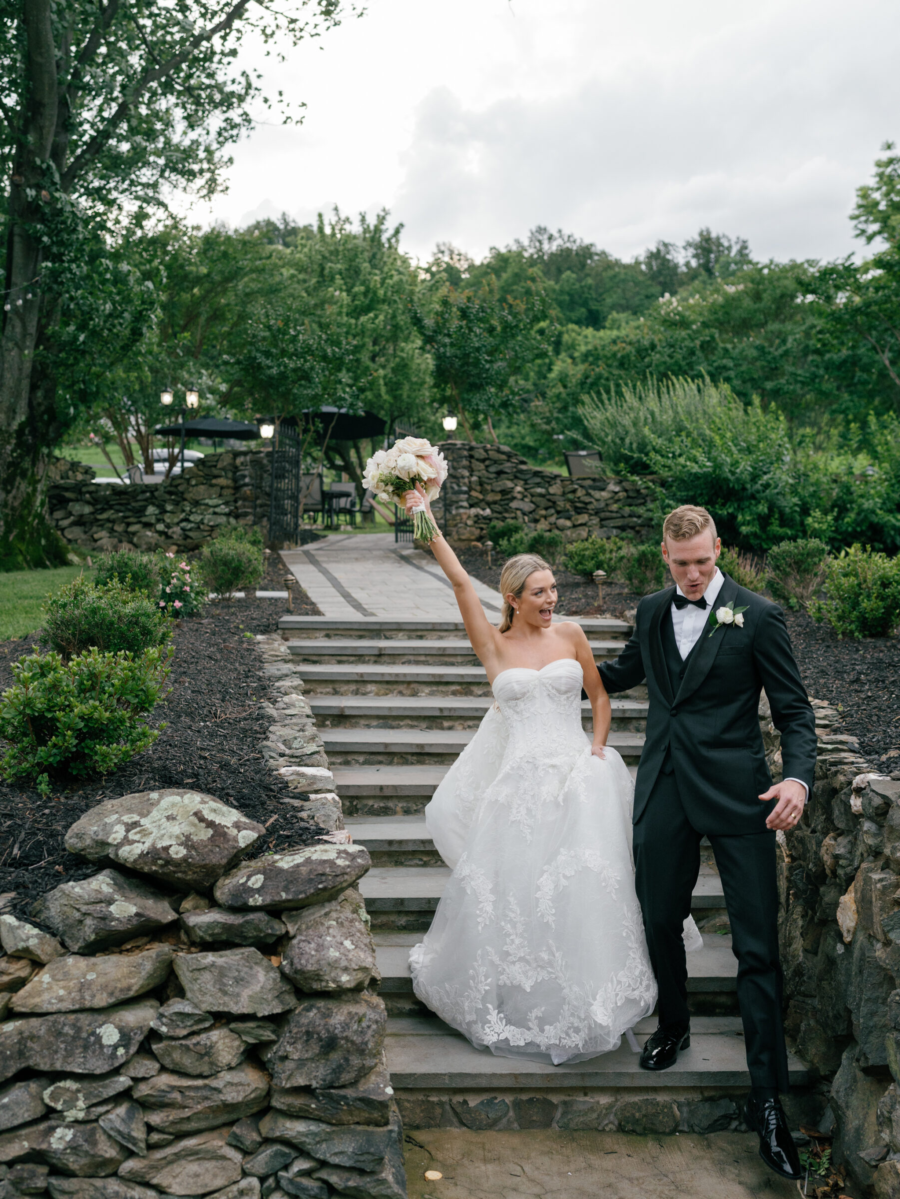 Newlyweds laugh together as they walk down the stone steps at The View at Bluemont, the bride raising her bouquet in joyful celebration.