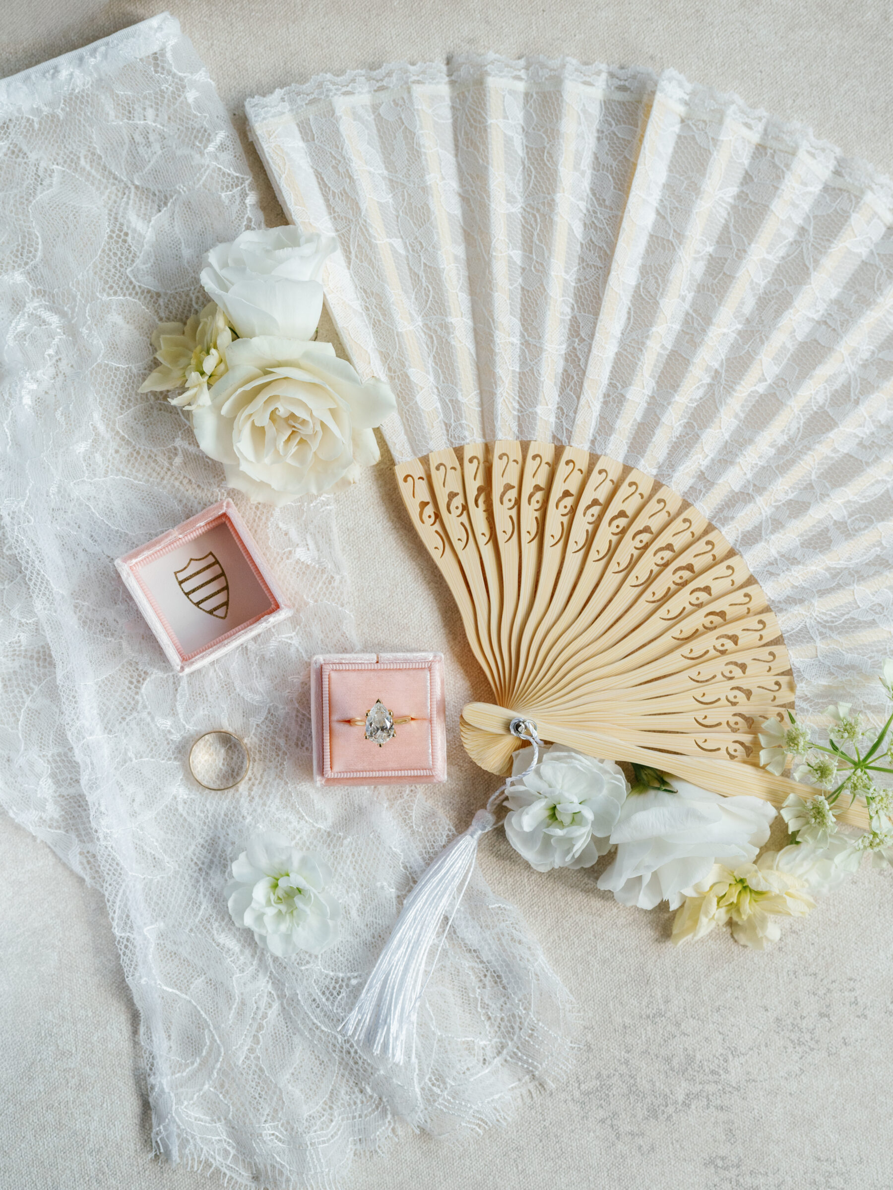 Elegant bridal flat lay featuring a lace hand fan, white roses, a pear-shaped engagement ring in a pink velvet box, and soft floral details.