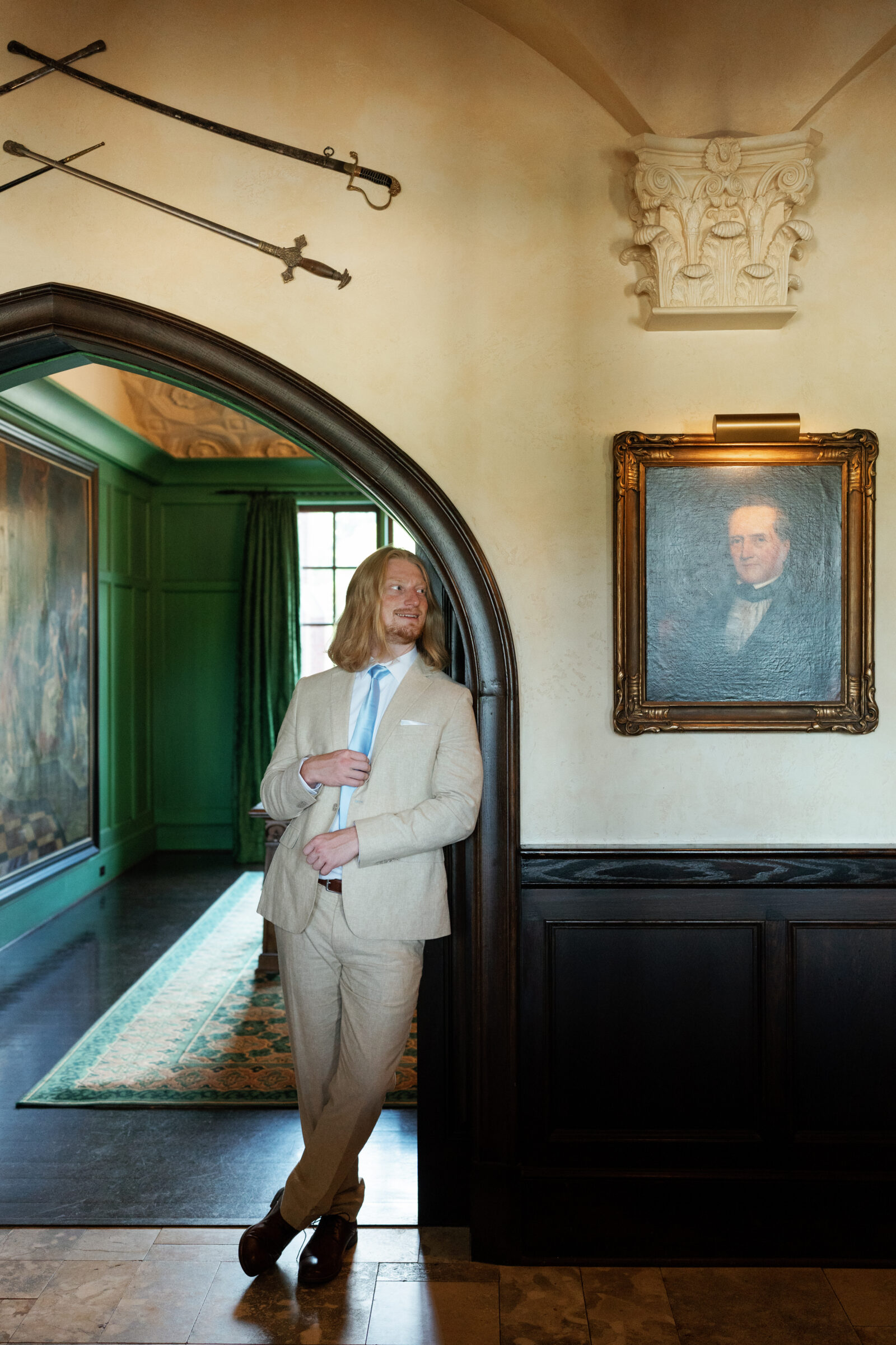 Groom in a khaki suit adjusting his tie inside Dover Hall, standing beneath an arched doorway surrounded by rich green walls and antique portraits — captured by a Richmond wedding photographer.