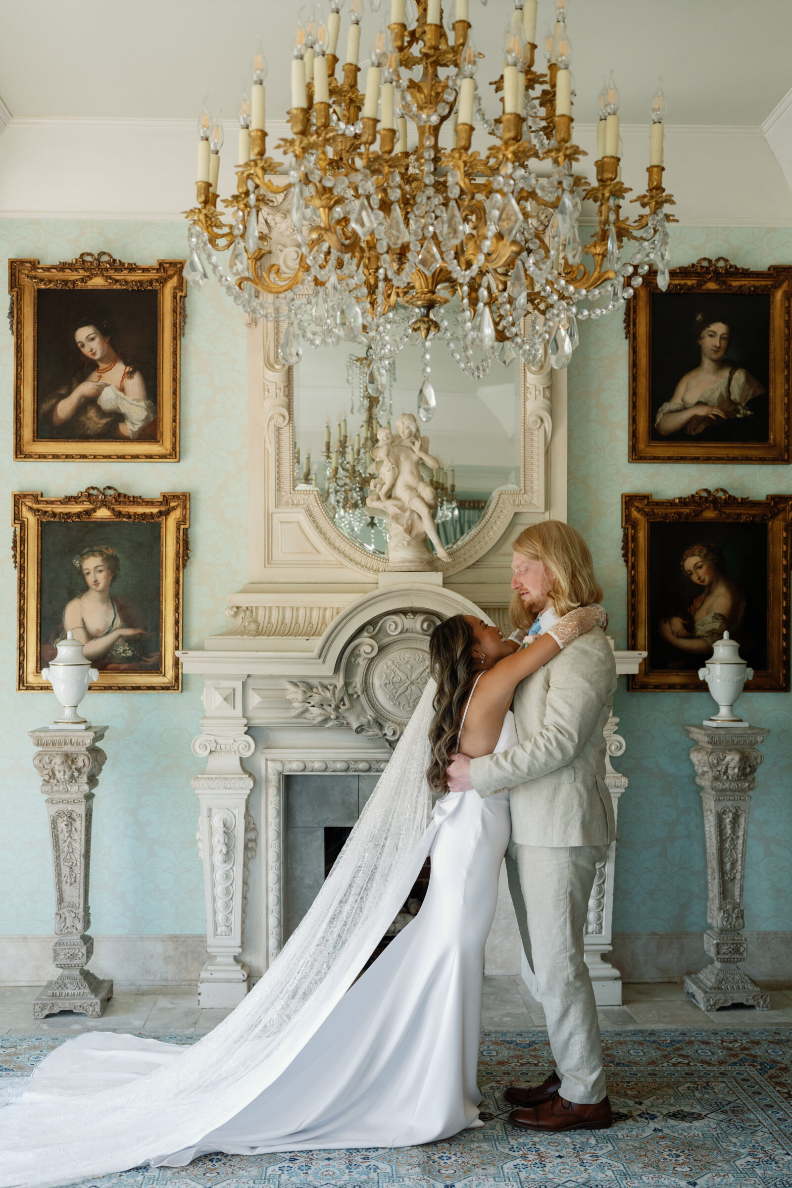 Bride and groom embrace in front of an ornate fireplace and chandelier inside Dover Hall, surrounded by classic paintings and pastel tones — a romantic moment captured by a Richmond wedding photographer during a Dover Hall wedding.
